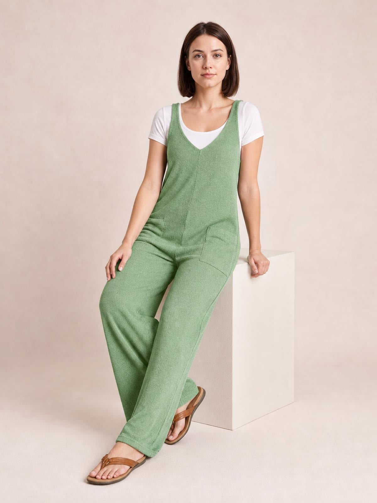 Model seated in a sage green textured sleeveless jumpsuit with a V-neck, wide-leg silhouette, front seam, and visible side pockets.