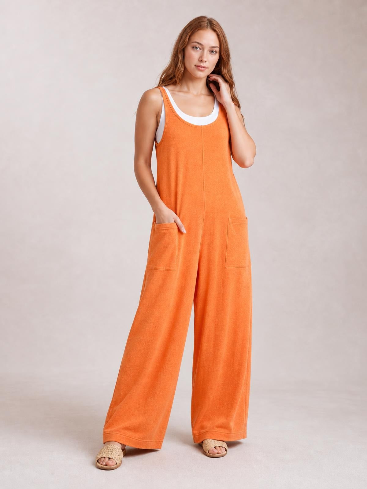 Model wears an orange textured sleeveless wide-leg jumpsuit with a relaxed silhouette, scoop neckline, front patch pockets, and one hand tucked inside.