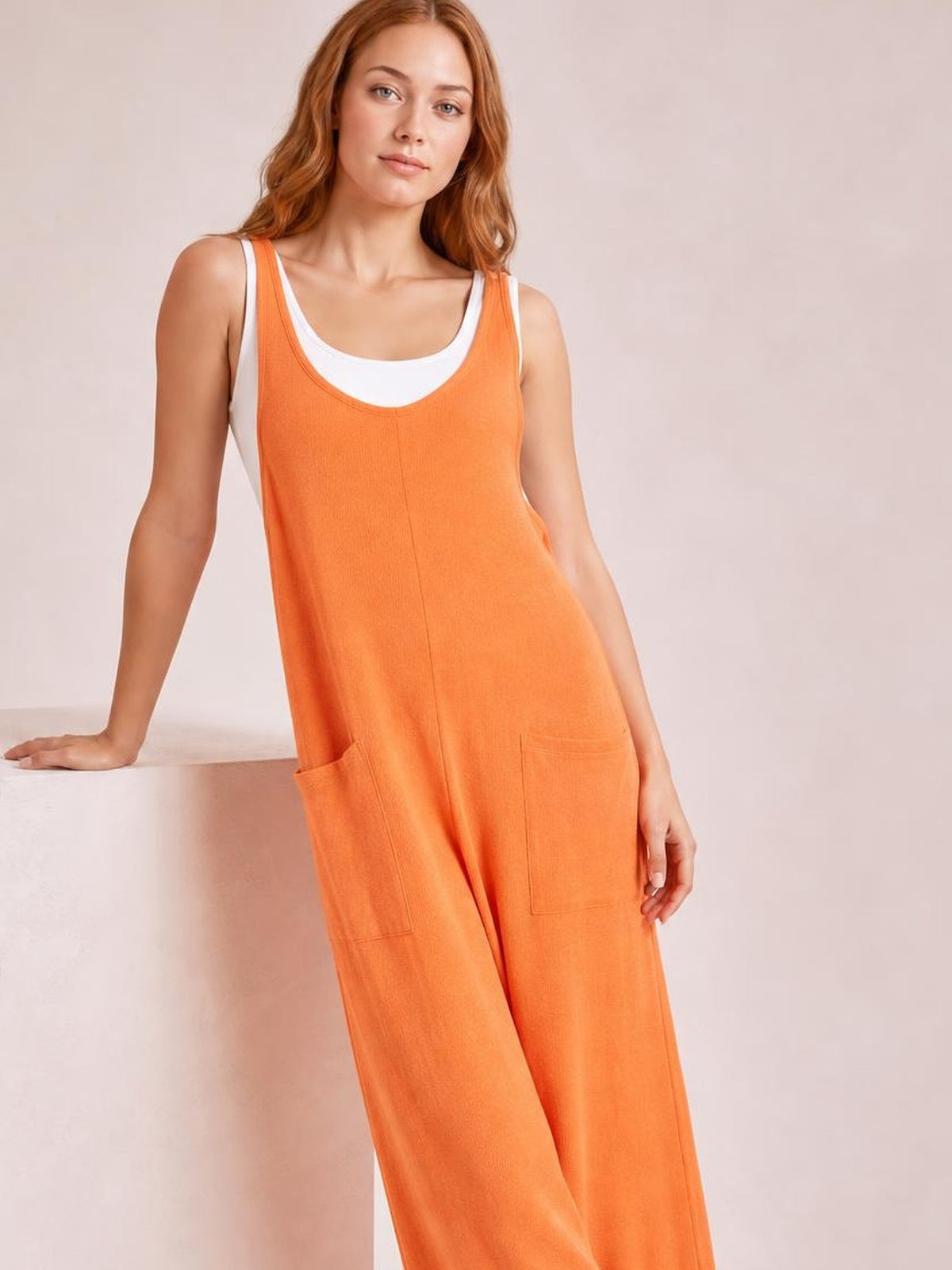 A model leans against a ledge wearing an orange textured sleeveless jumpsuit with a relaxed silhouette, scoop neckline, and front patch pockets.