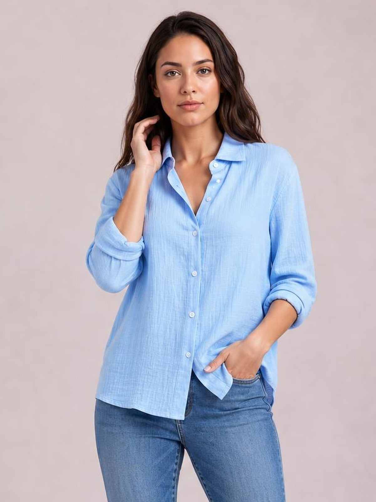 A model wears a light blue textured button-front shirt with a collared neckline, long rolled sleeves, and a relaxed curved hem, standing with one hand in her pocket.