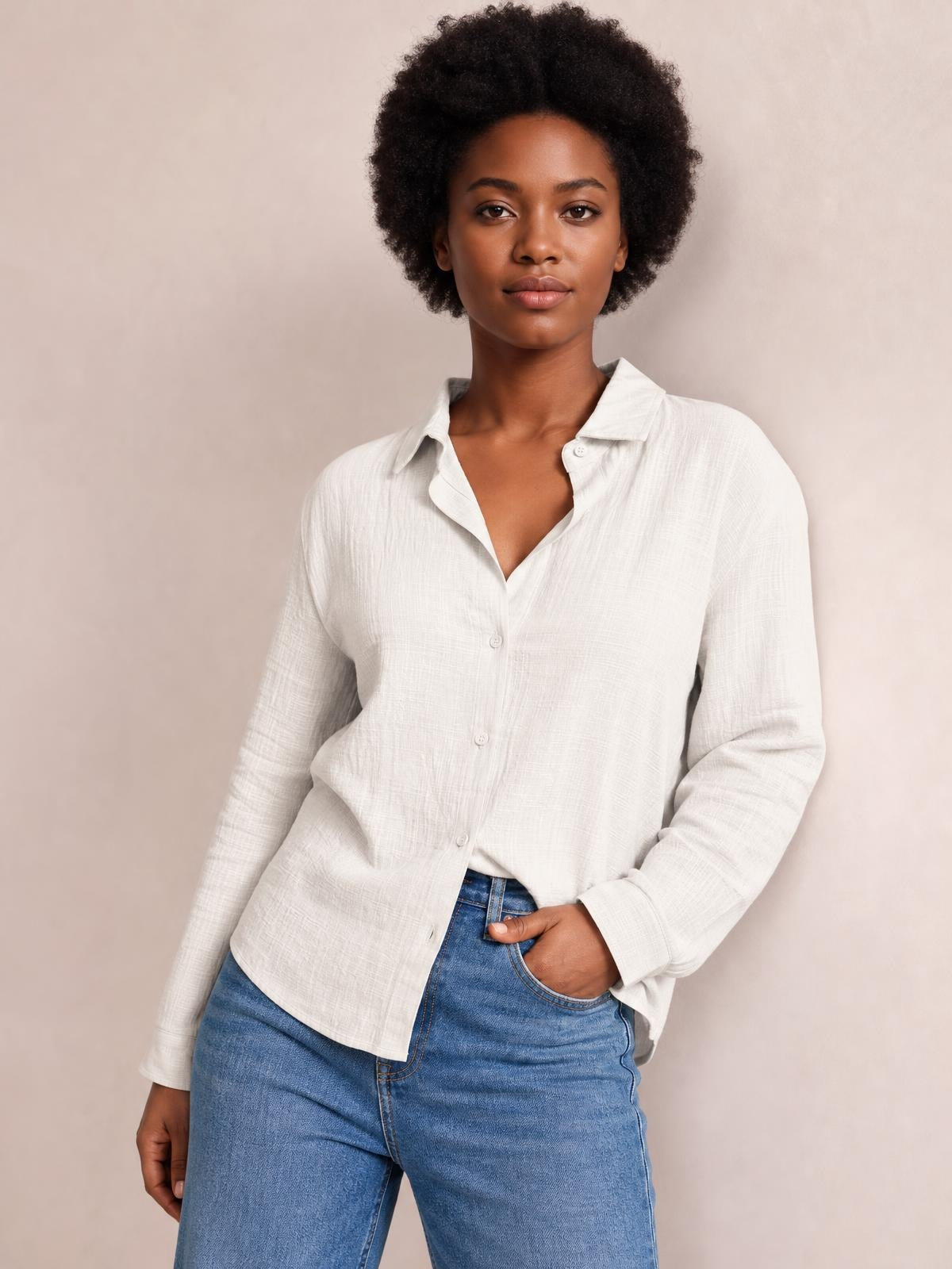 Model wears a white textured button-front shirt with a collared neckline, long sleeves, relaxed silhouette, and curved hem, posing with one hand in her jeans pocket.