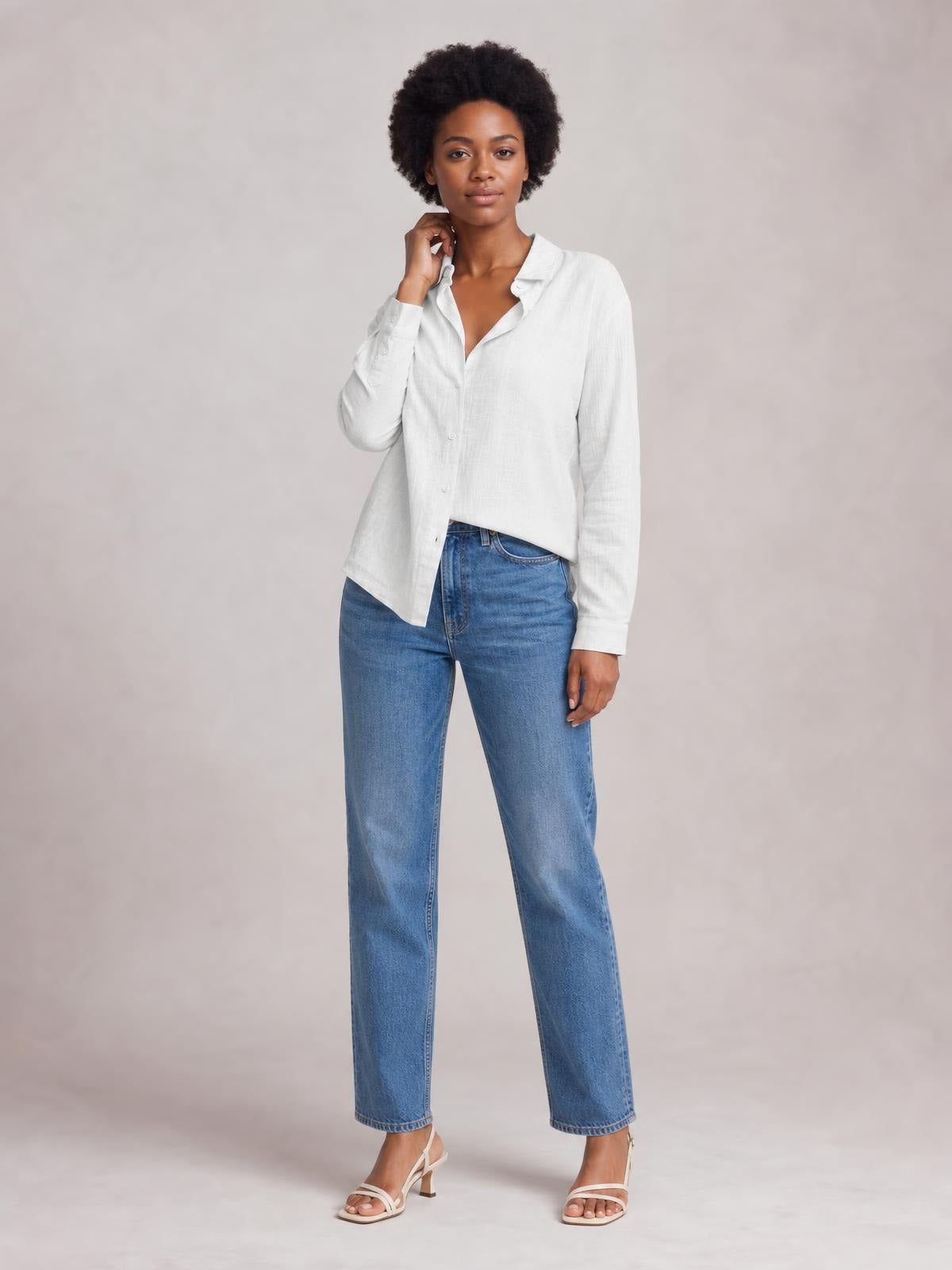 Model wearing a white textured button-front shirt with a collared neckline, long sleeves, relaxed silhouette, and curved hem, posing with one hand near her neck.