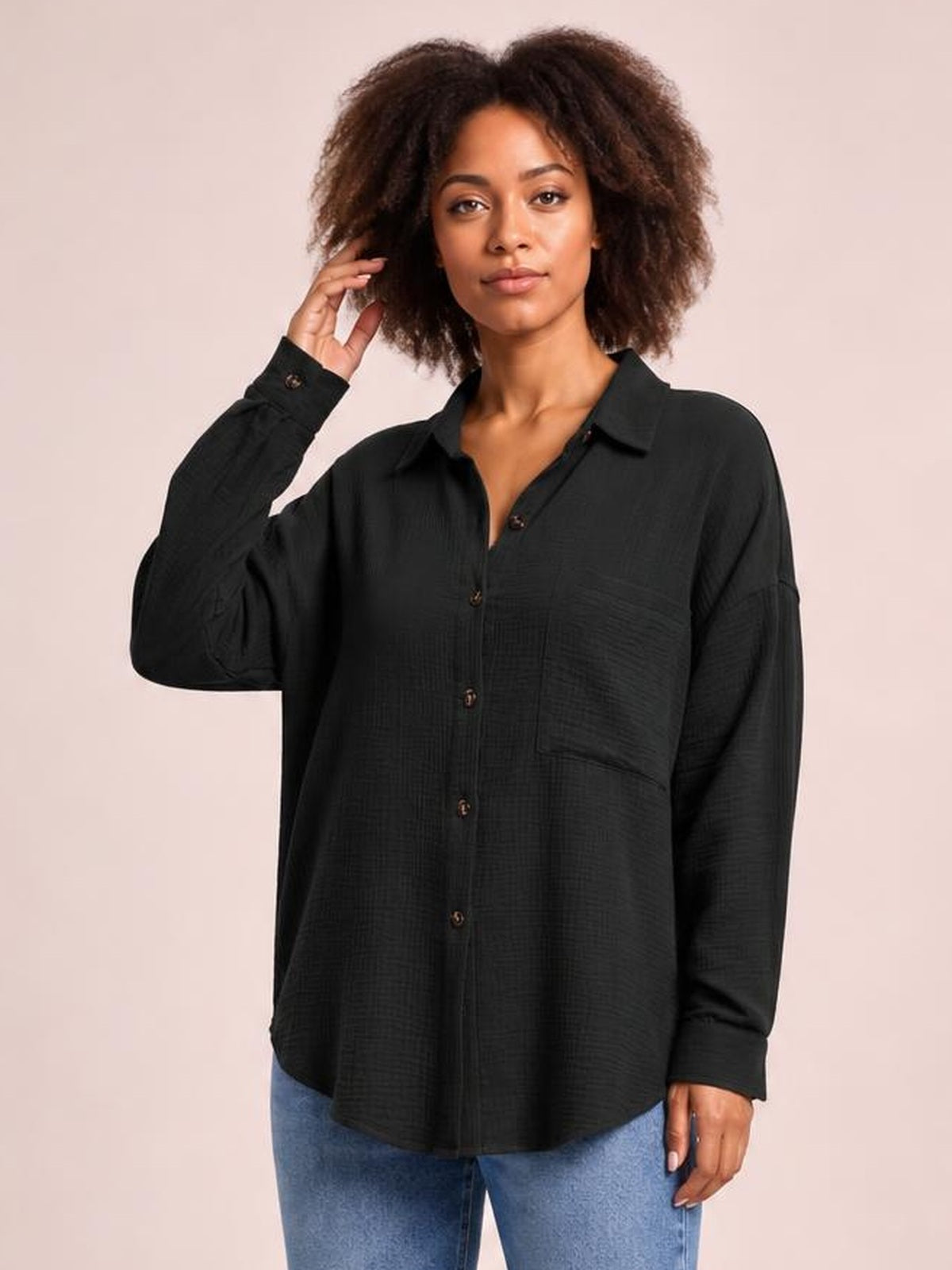 Model wears a black textured longline shirt with a collared neckline, button front, dropped shoulders, long sleeves, and a chest pocket while touching her hair.