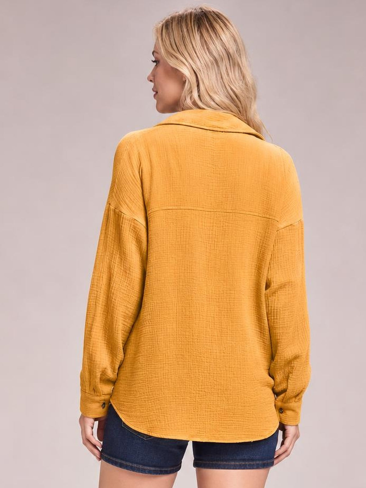 A model faces away wearing a mustard textured longline shirt with a collared neckline, dropped shoulders, long sleeves, and a relaxed rounded-hem silhouette.