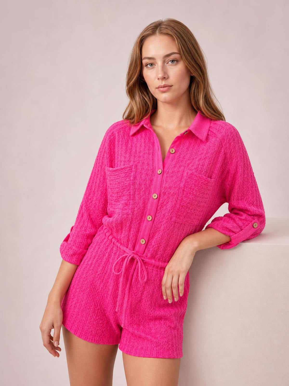 A model poses in a bright pink textured knit button-front romper with a collared neckline, long sleeves, patch pockets, drawstring waist, and relaxed shorts silhouette.