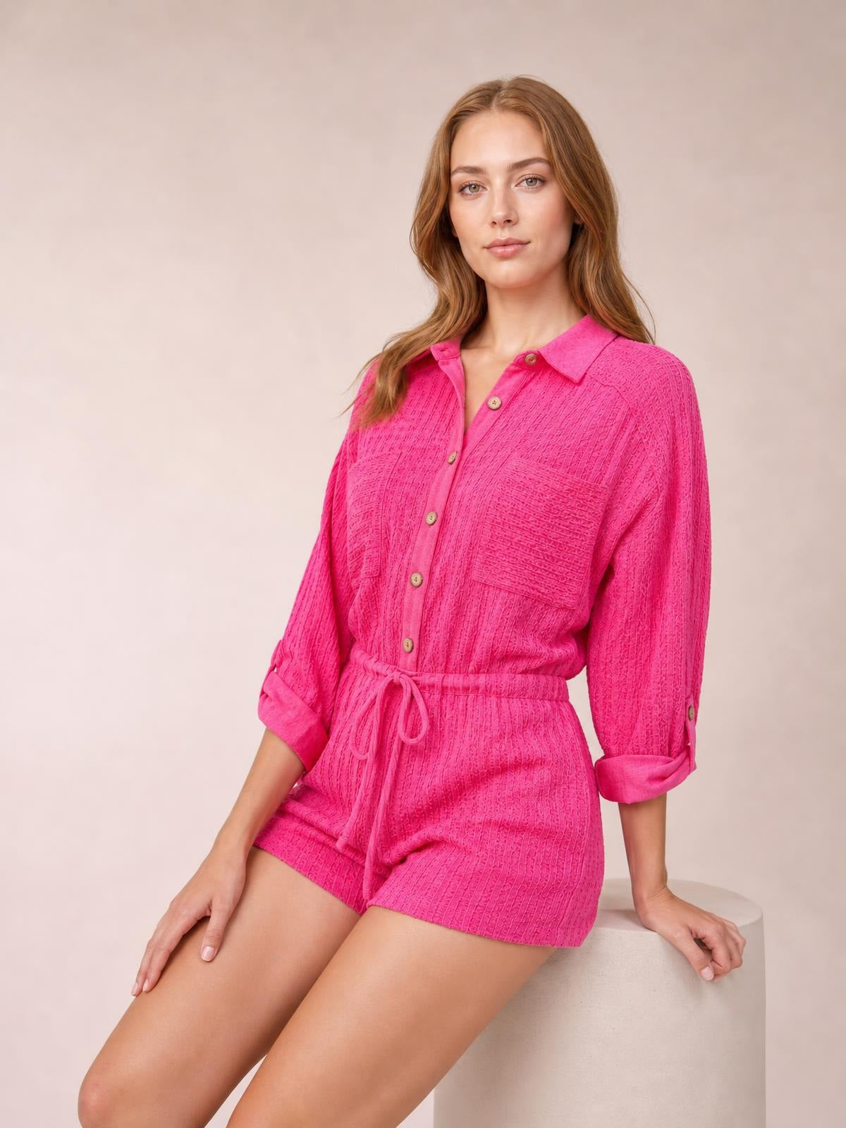 Model seated in a bright pink textured knit romper with a button-front shirt collar, long rolled sleeves, drawstring waist, and relaxed shorts silhouette.