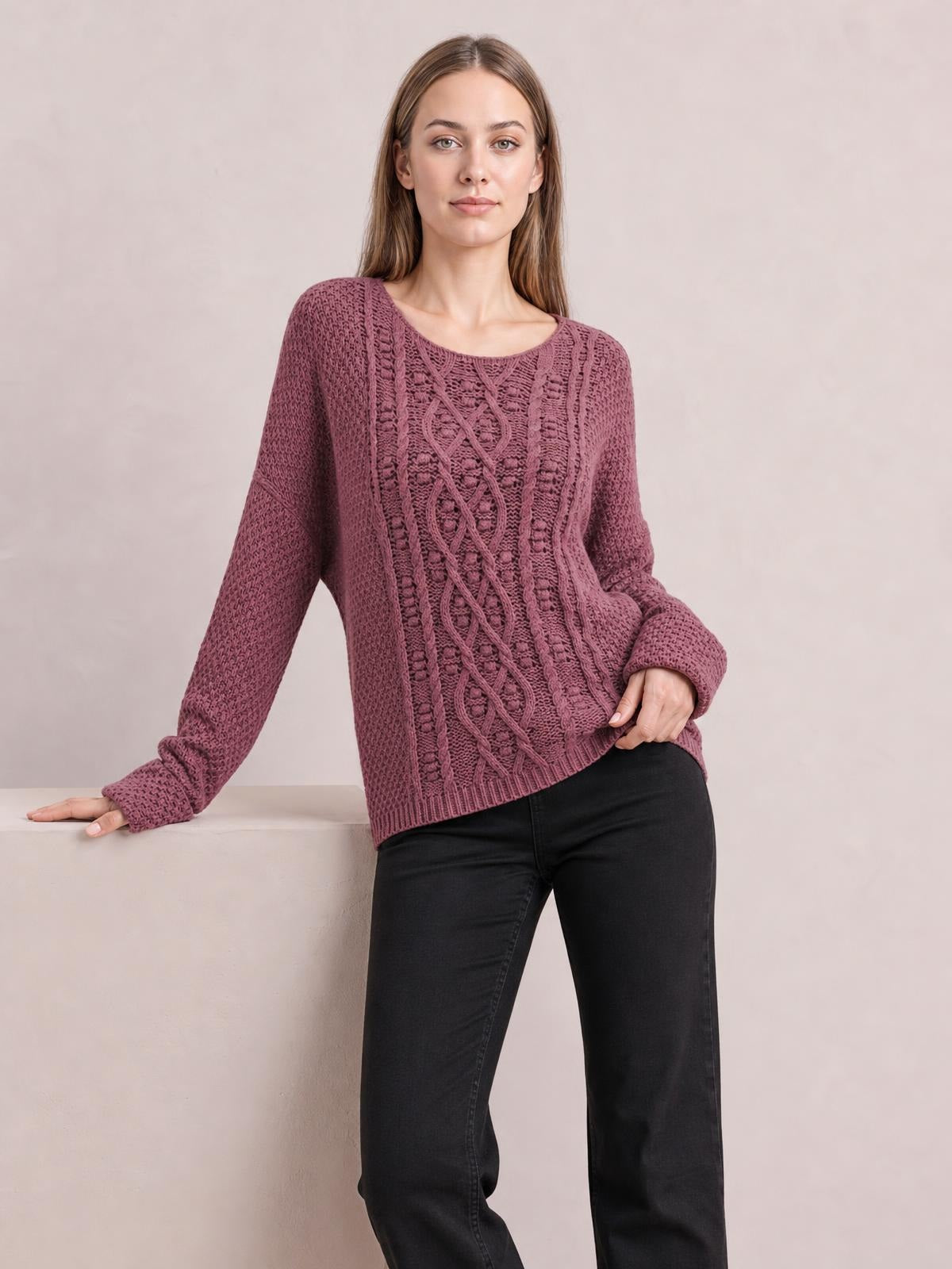 Model wears a mauve textured cable-knit round-neck sweater with a relaxed straight silhouette, long sleeves, ribbed cuffs and hem, posing with one hand on a ledge.