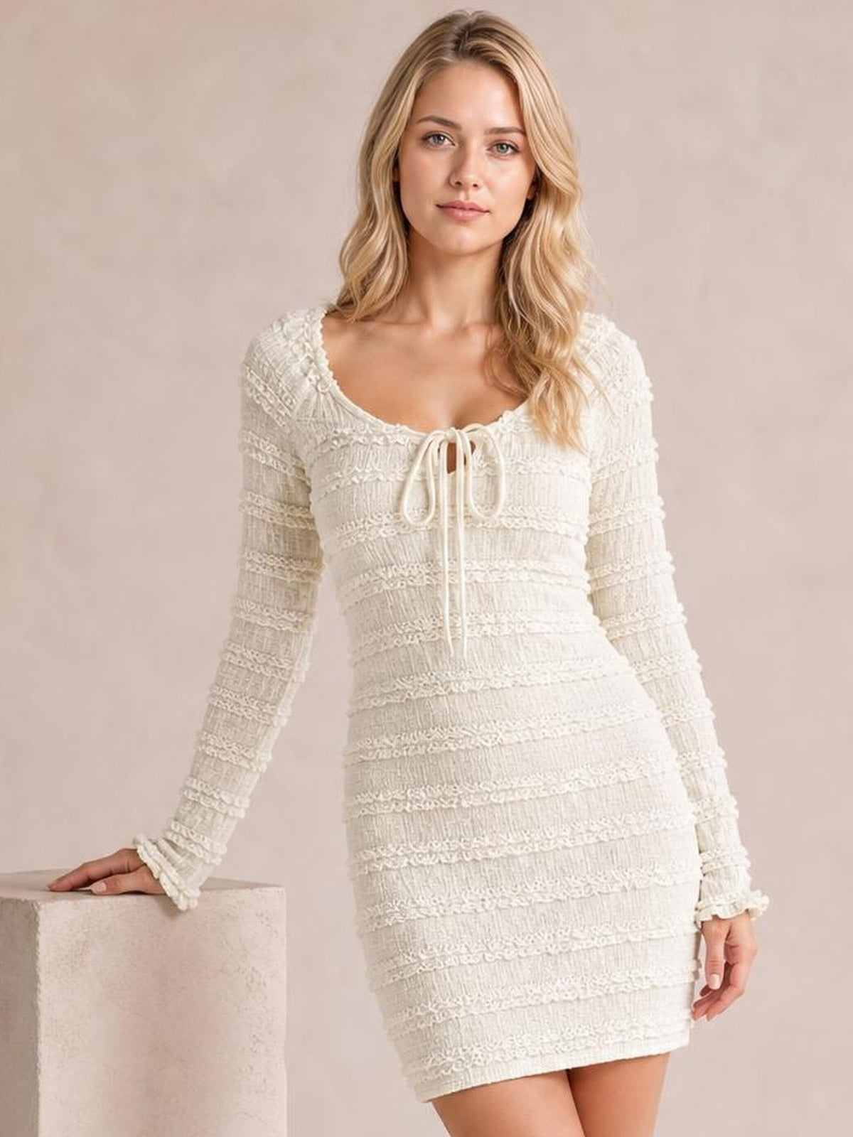 Model poses with one hand on a block in a cream textured long-sleeve bodycon mini dress featuring a scoop neckline, tie-front, and fitted silhouette.