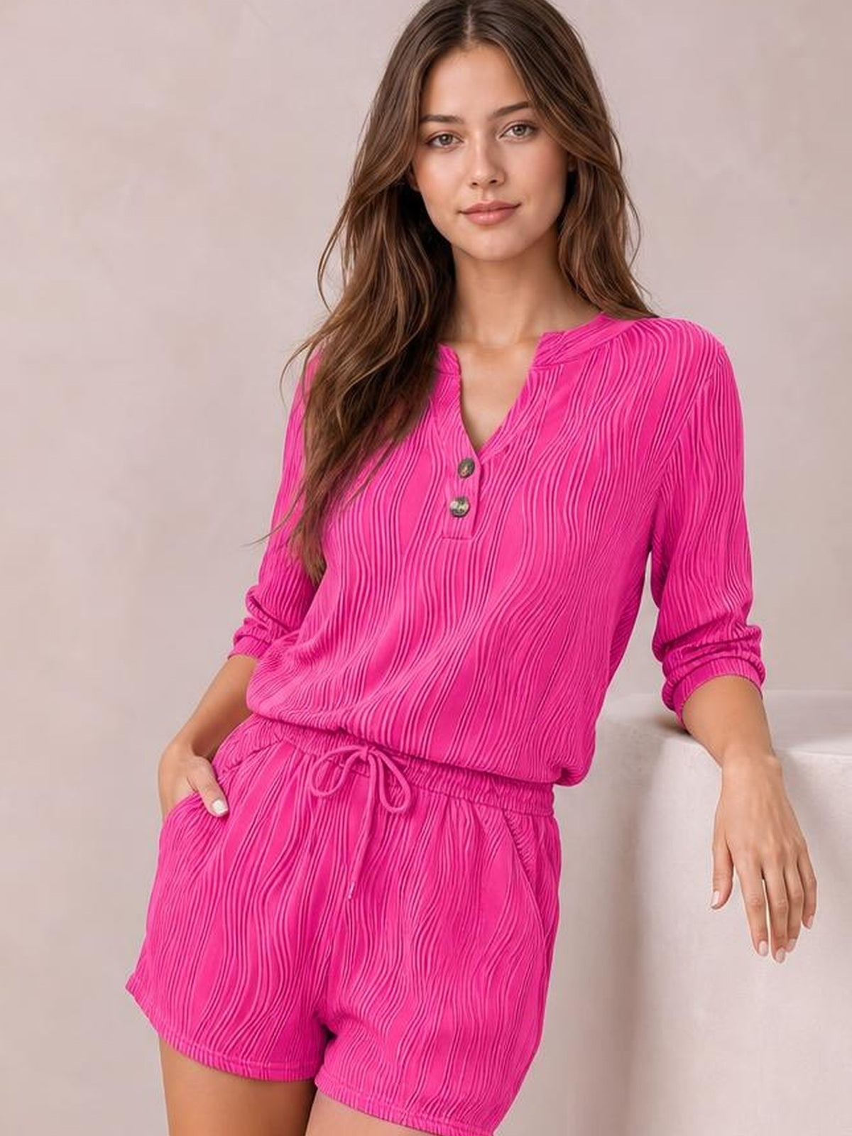 A seated model wears a bright pink textured notched top with button detail and rolled sleeves, paired with drawstring shorts featuring an elastic waist and pockets.
