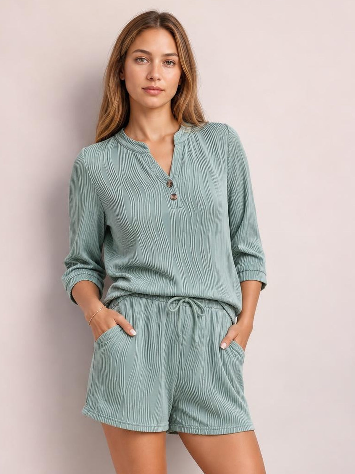 Model wears a sage green textured notched top with button placket and long sleeves, paired with relaxed drawstring shorts featuring pockets and a fluid silhouette.