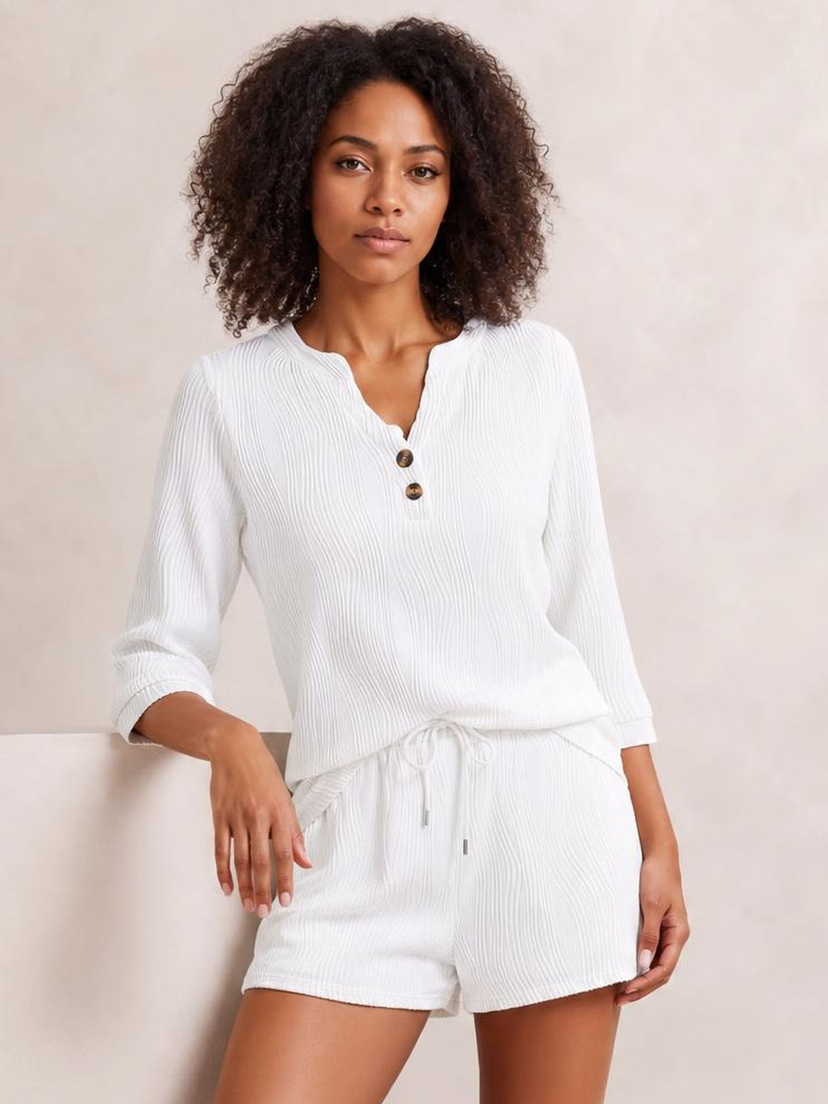 Model poses in a white textured notched top with long sleeves and button detail, paired with relaxed drawstring shorts for a comfortable set silhouette.