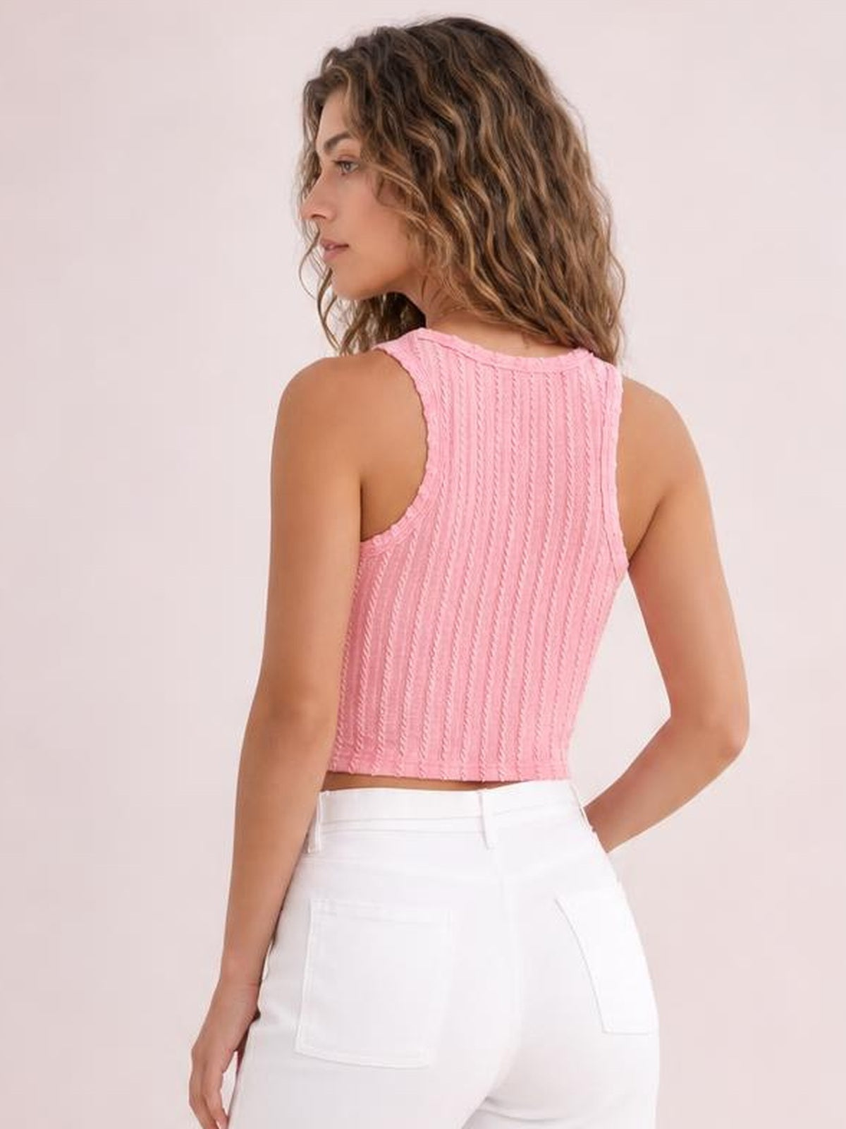 A model poses with one hand on her hip, showing the pink textured racerback cropped tank’s fitted silhouette, high neckline, and vertical knit-like construction.