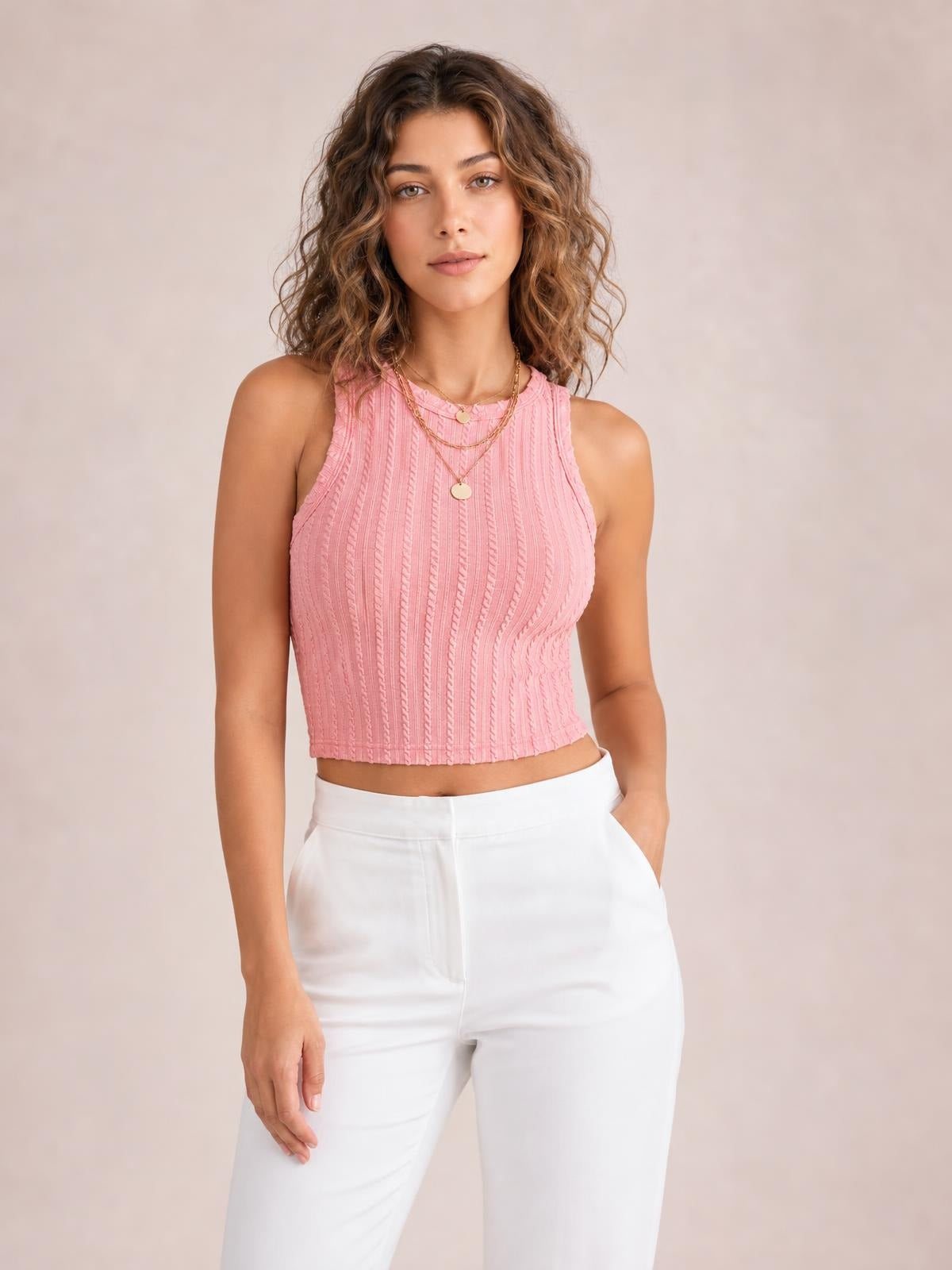 A model stands in a pink textured cropped tank with a high neckline, sleeveless cut, fitted silhouette, and vertical detailing, paired with white pants and one hand in a pocket.
