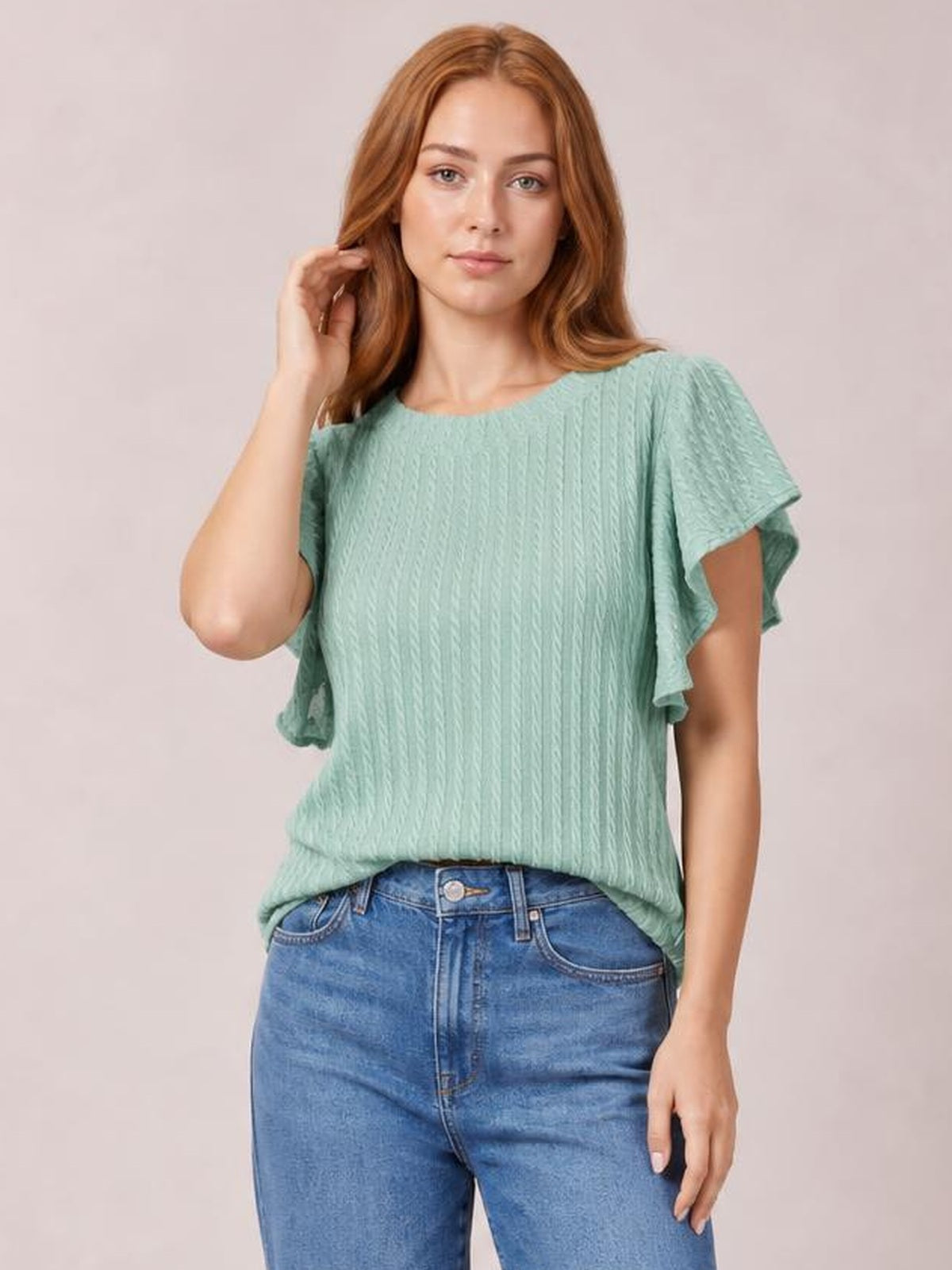 A model touches her hair while wearing a mint green textured round-neck blouse with layered flounce sleeves and a relaxed straight silhouette.