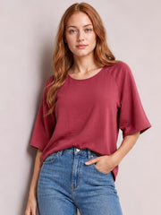 Model wearing a solid burgundy textured round-neck top with relaxed long silhouette, half cold-shoulder sleeves, and a casual fit, posing with one hand in her jeans pocket.