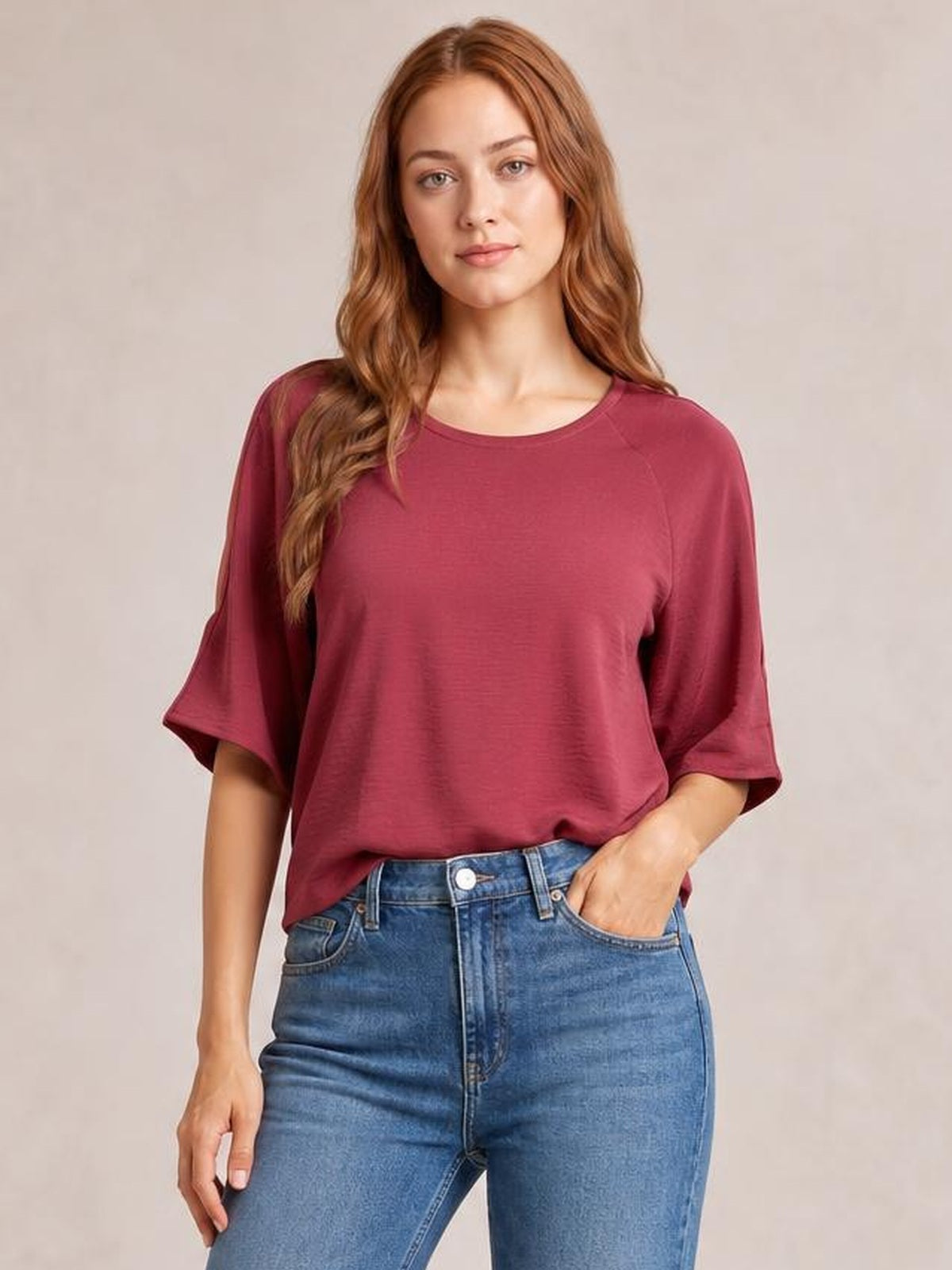 A model wears a deep burgundy textured round-neck top with a relaxed long silhouette and wide split half sleeves, posing with one hand in her jeans pocket.