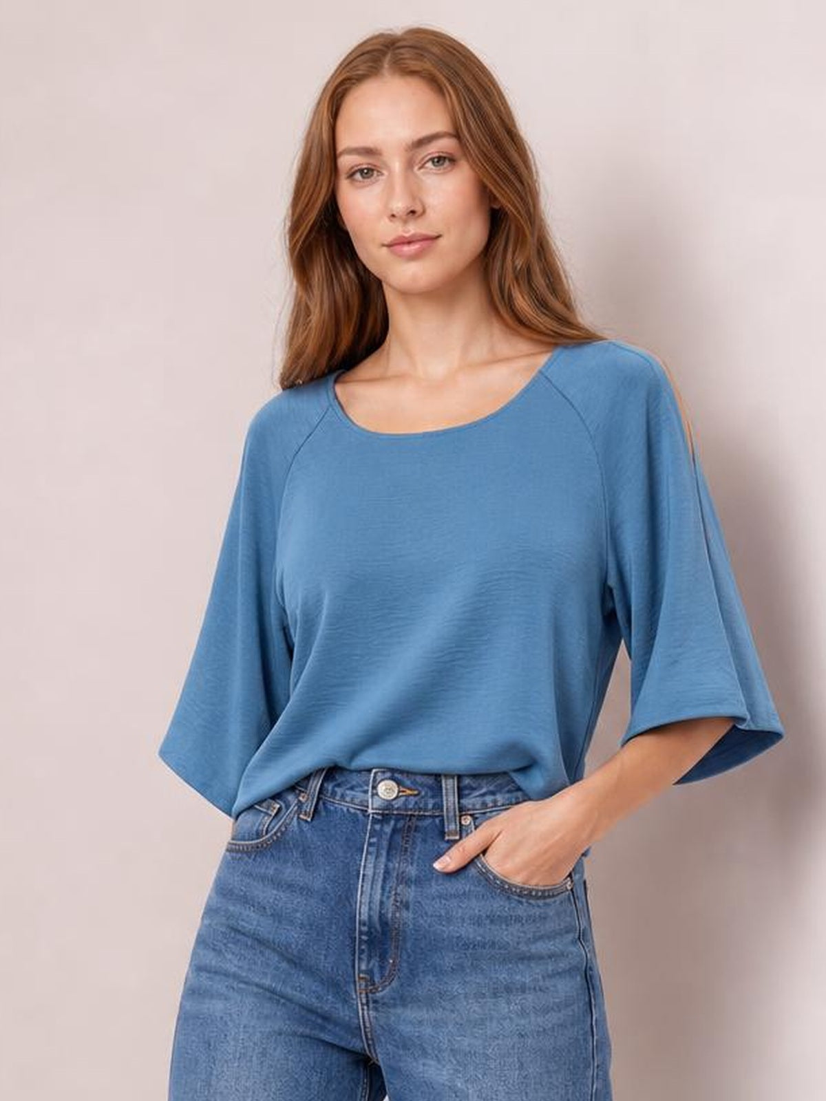 Model wearing a solid blue textured top with a round neckline, loose long silhouette, and wide split half sleeves, standing with one hand in her jeans pocket.