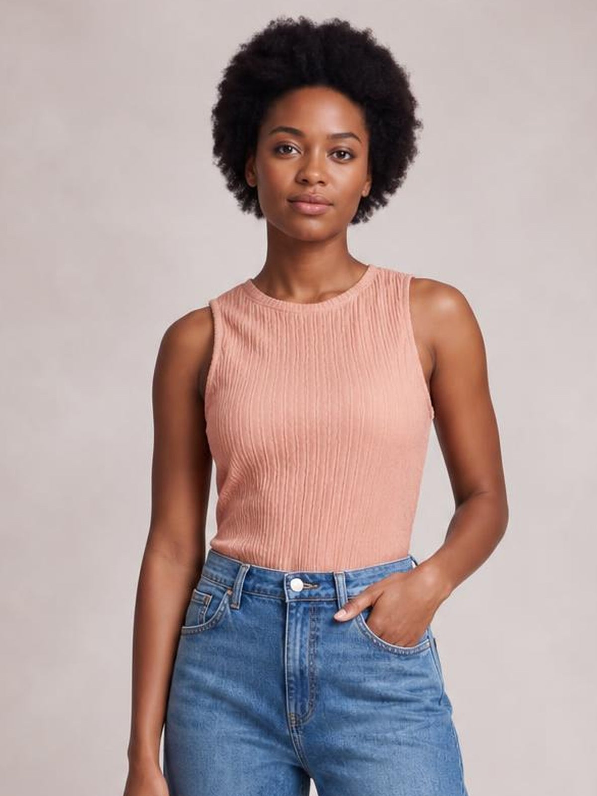 Model wearing a blush pink textured round neck tank with a sleeveless cut and fitted silhouette, standing with one hand tucked into her jeans pocket.