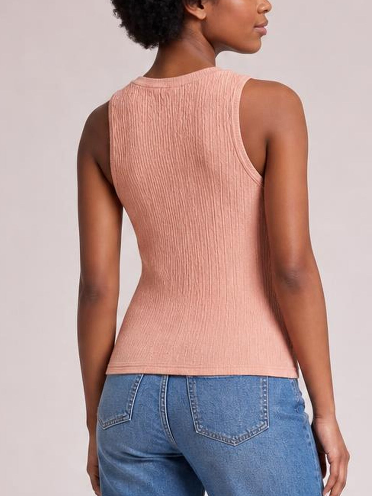A model faces away wearing a pink textured round neck tank with wide shoulder straps, a fitted silhouette, and neatly finished armholes and hem.