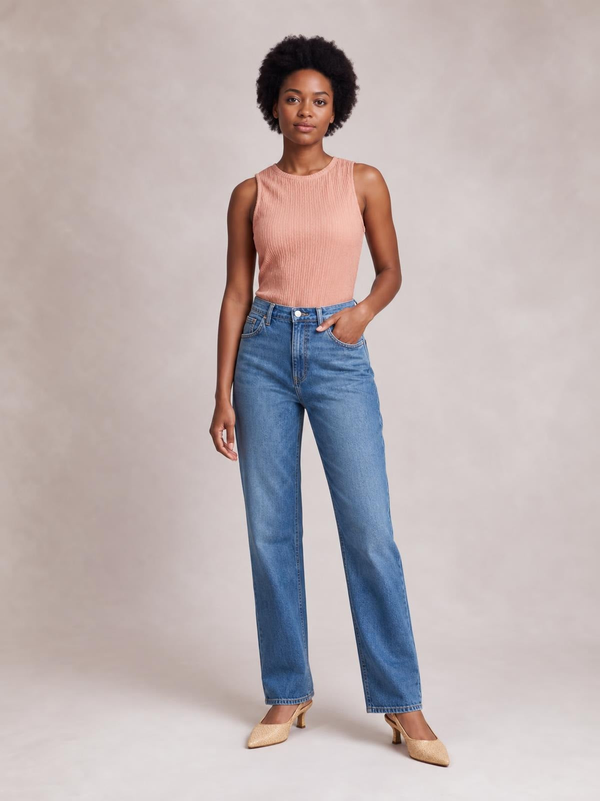 Model wears a blush pink textured round neck tank with a fitted sleeveless silhouette and ribbed construction, posed with one hand in her jeans pocket.