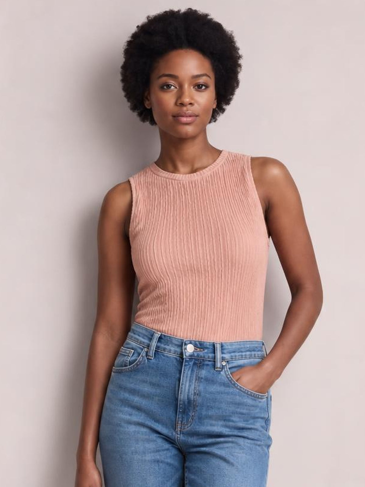 Model poses with one hand in her pocket wearing a blush pink textured round neck tank with vertical ribbing and a fitted sleeveless silhouette.