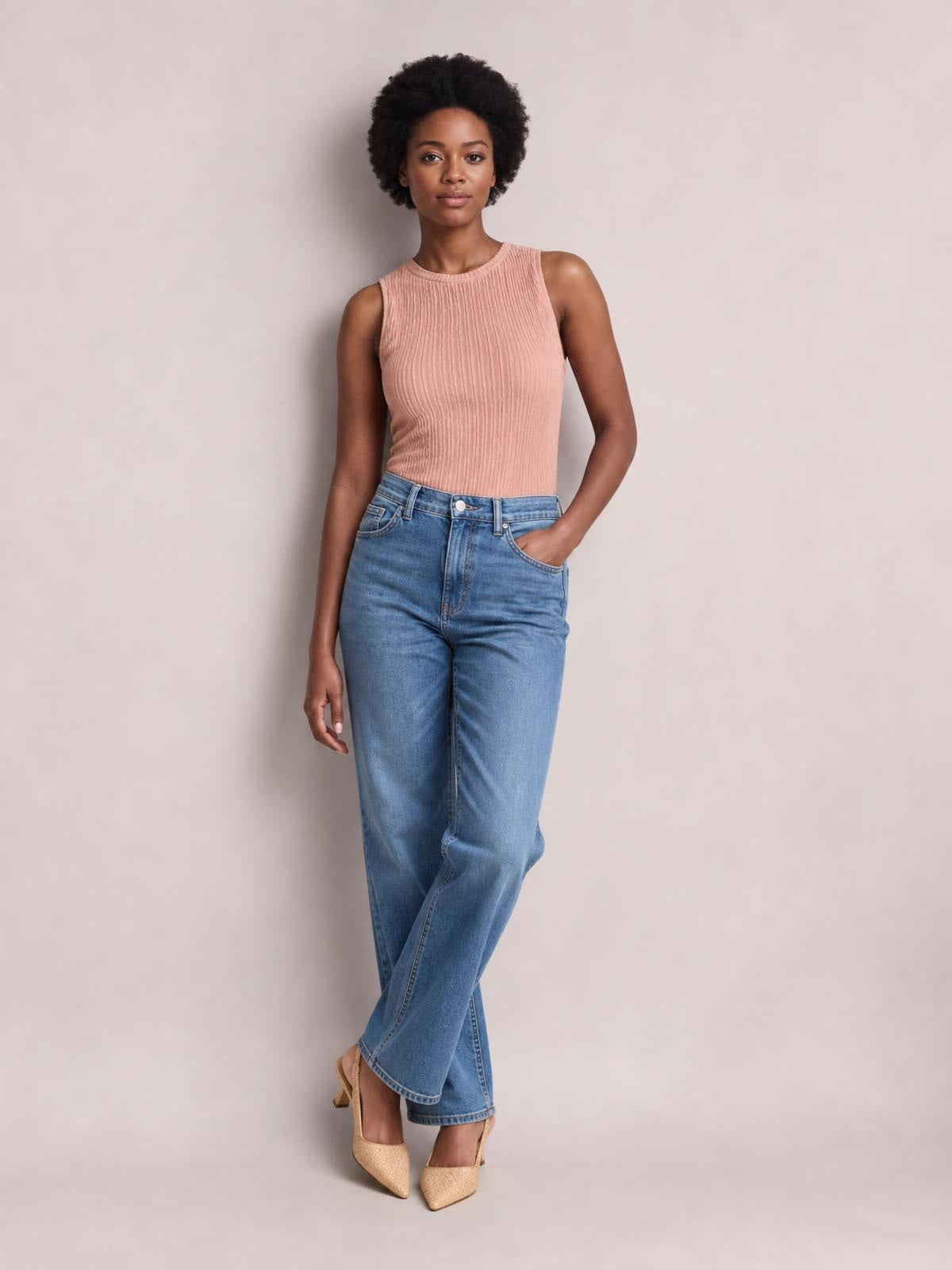 Model wears a blush pink textured round neck tank with a sleeveless, fitted silhouette, standing with one hand in her pocket and crossed legs.