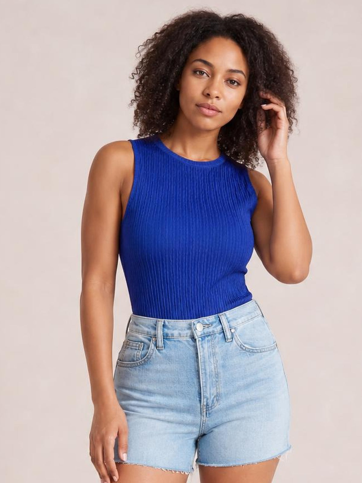 Model wearing a royal blue textured round neck tank with a fitted, slightly stretchy silhouette, standing with one hand touching her hair.