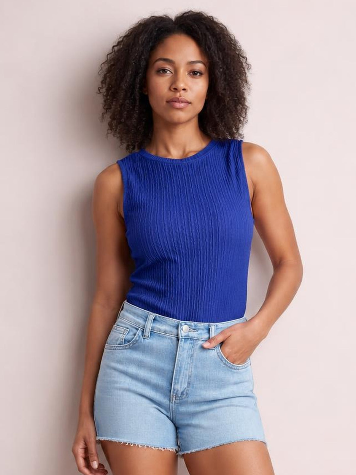 Model wearing a blue textured round neck tank with a sleeveless, fitted silhouette and vertical crinkled construction, paired with denim shorts.