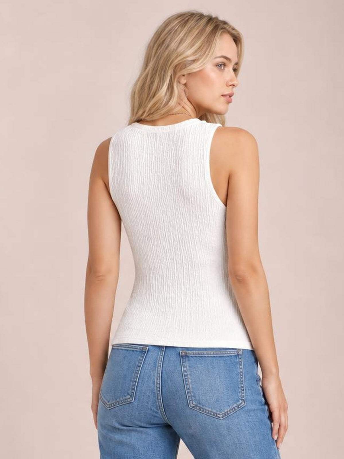 A model looks over her shoulder wearing a white textured round neck tank with a sleeveless cut and fitted silhouette, paired with blue jeans.