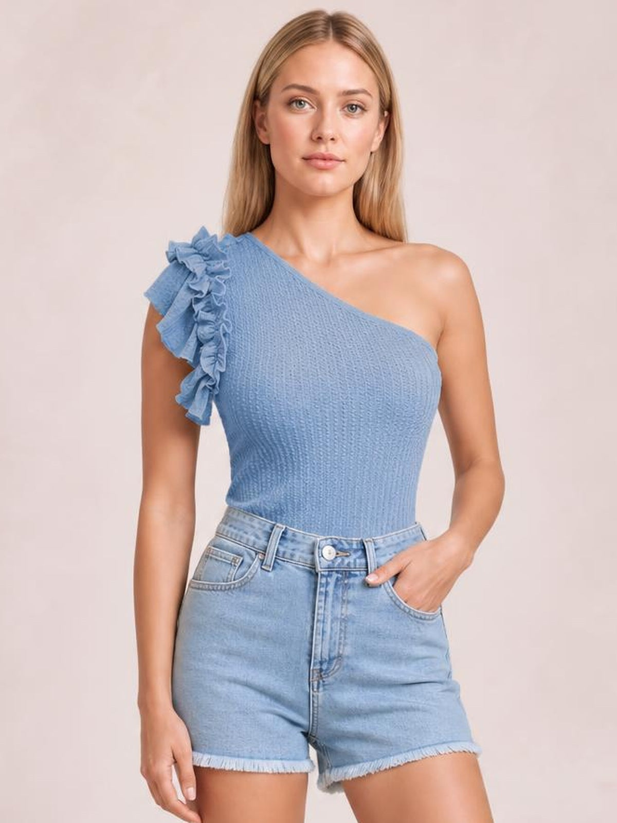 Model wearing a blue textured one-shoulder top with layered ruffled trim and a fitted silhouette, paired with light-wash denim shorts.