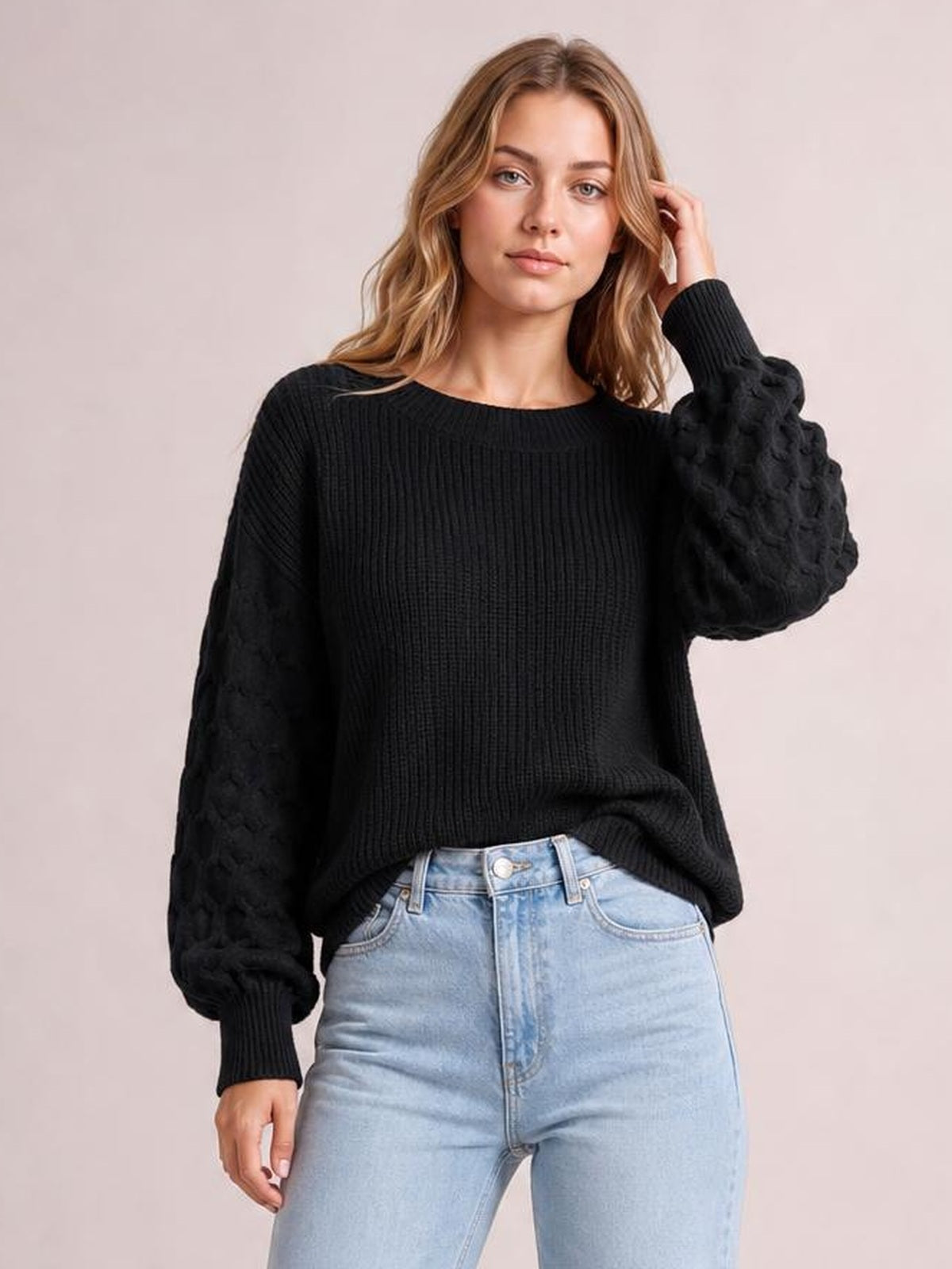 Model wearing a black textured sleeve round neck sweater with ribbed cuffs and hem, relaxed silhouette, standing with one hand touching her hair.