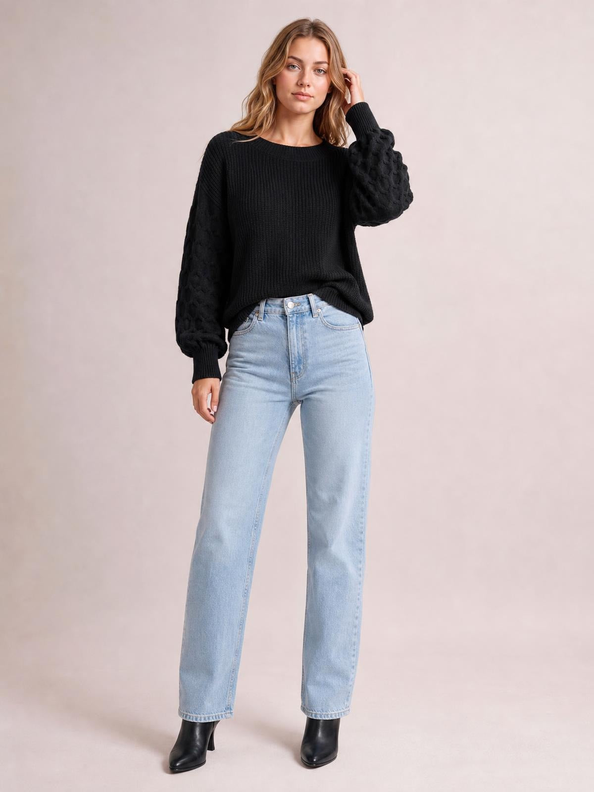 Model wearing a black textured sleeve round neck sweater with a ribbed relaxed silhouette, standing in light-wash straight-leg jeans with one hand touching her hair.