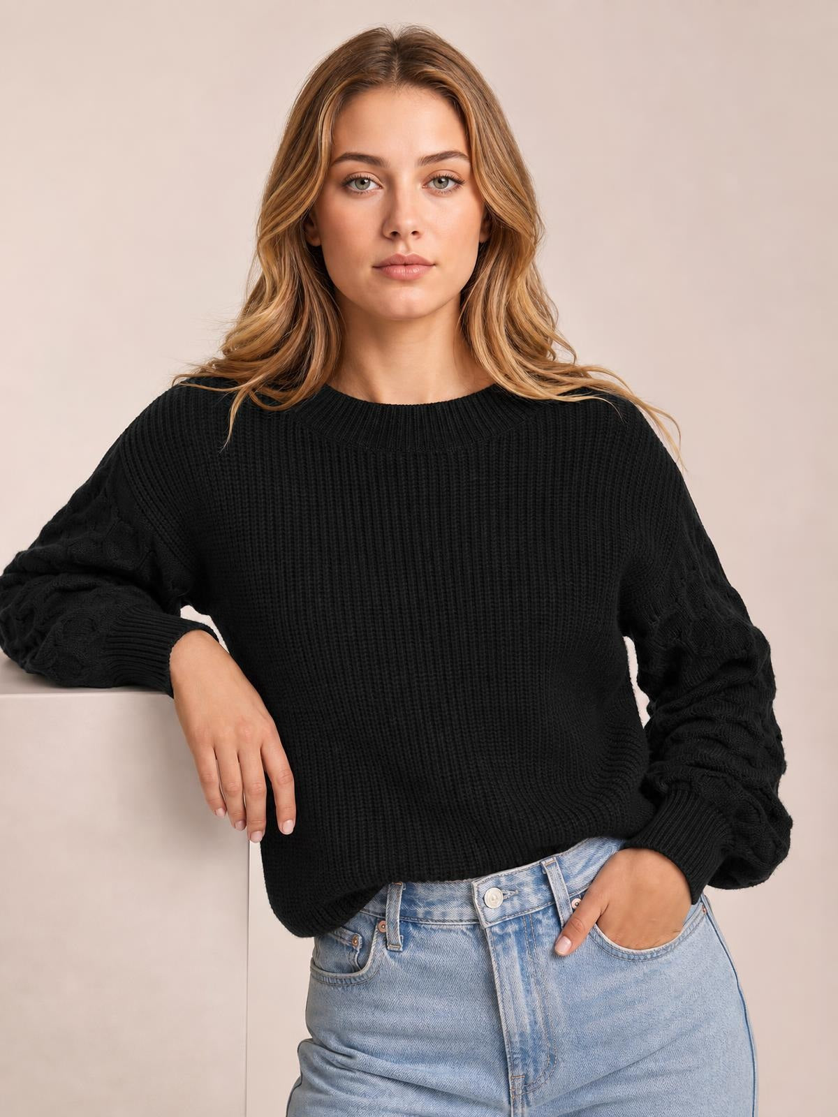Model wearing a black ribbed round neck sweater with textured balloon sleeves and a relaxed silhouette, leaning on a surface with one hand in her jeans pocket.
