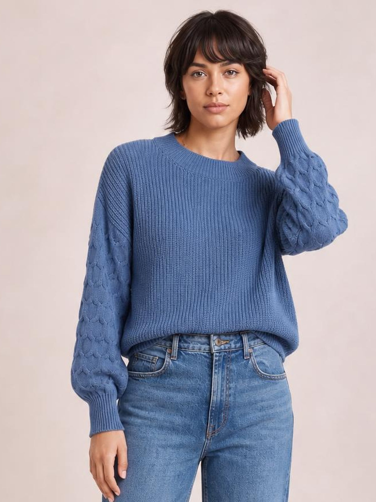 Model wearing a blue textured sleeve round neck sweater with ribbed cuffs and hem, a relaxed silhouette, and one hand raised to her hair.