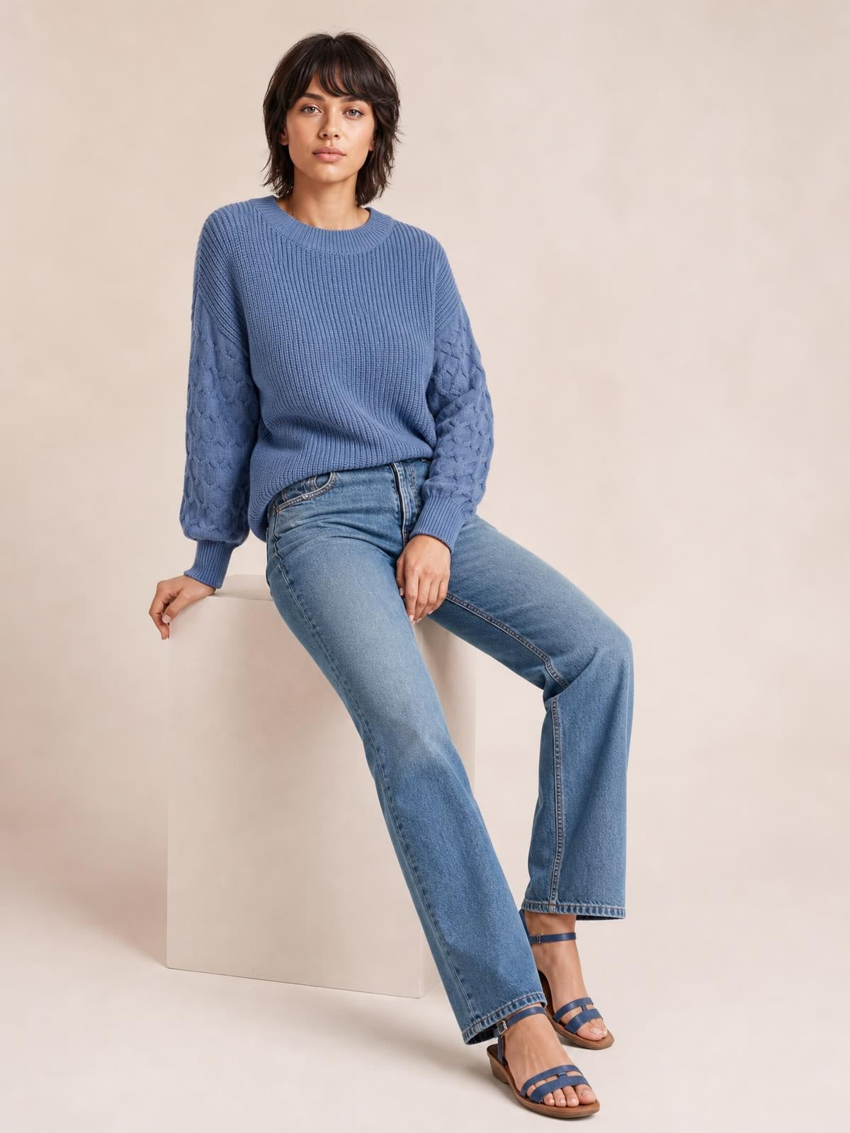 A model sits on a block wearing a blue textured sleeve round neck sweater with ribbed trim, patterned sleeves, and a relaxed silhouette.