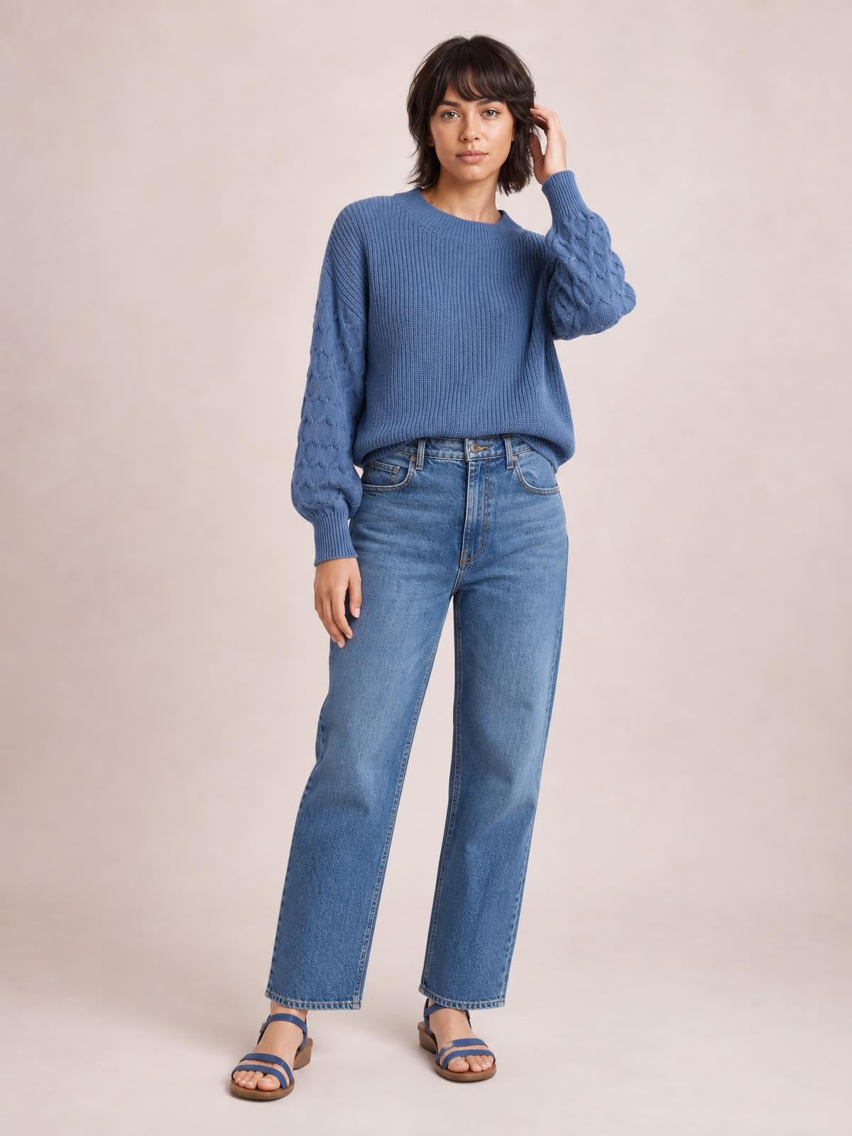 Model touches her hair while wearing a blue textured sleeve round neck sweater with ribbed knit construction, cable-textured sleeves, and a relaxed silhouette.