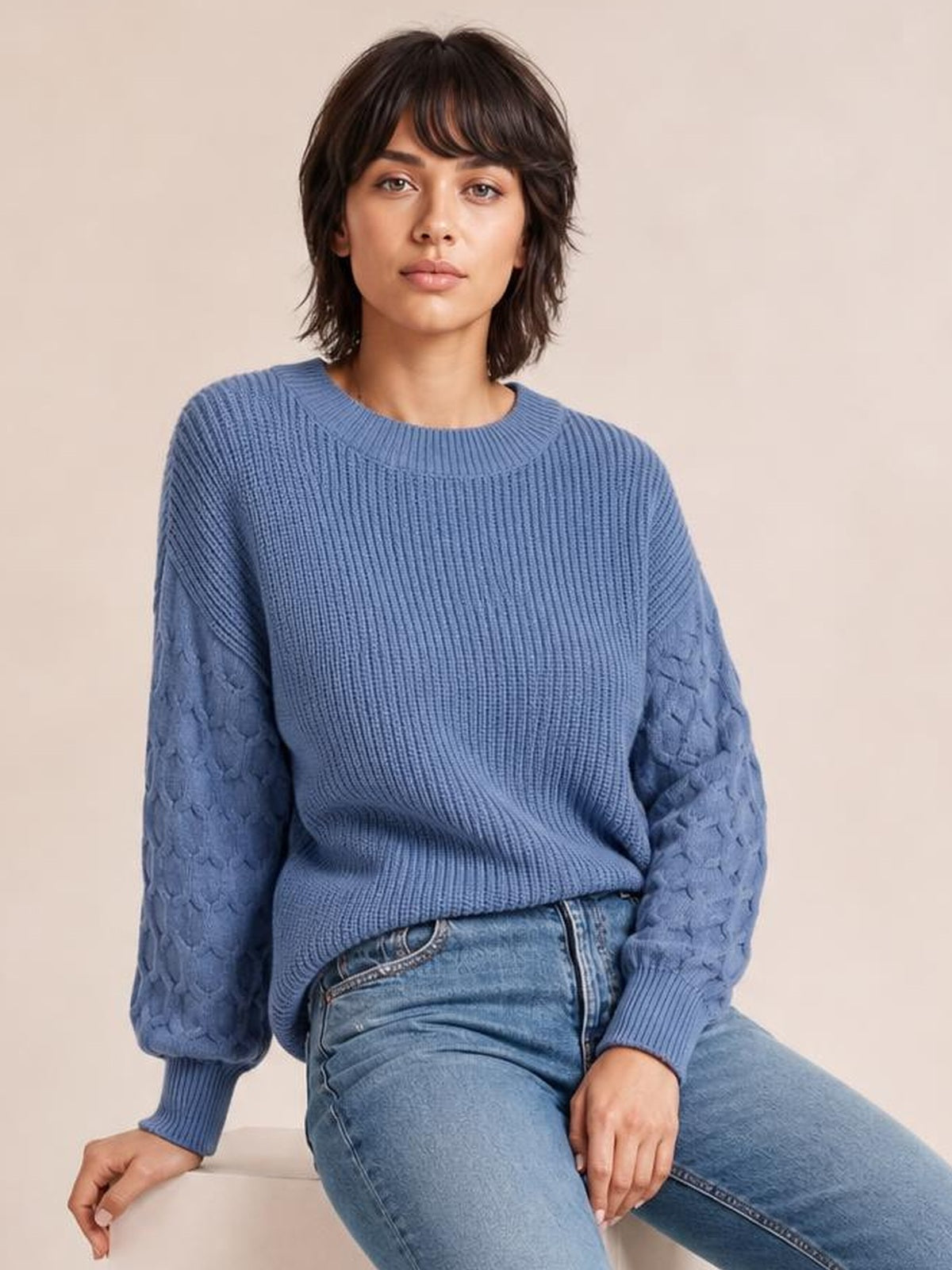Model seated in a blue textured sleeve round neck sweater with a ribbed knit body, patterned sleeves, relaxed silhouette, and ribbed cuffs.