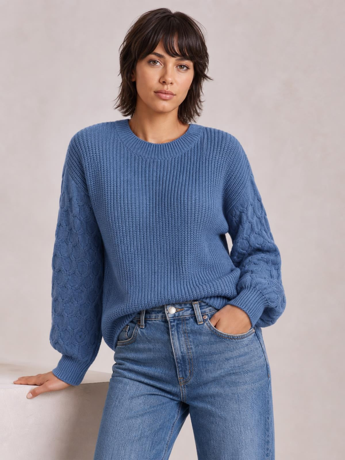 Model poses in a blue round-neck sweater with a ribbed body, textured patterned sleeves, relaxed silhouette, and ribbed cuffs, one hand resting and one in a pocket.