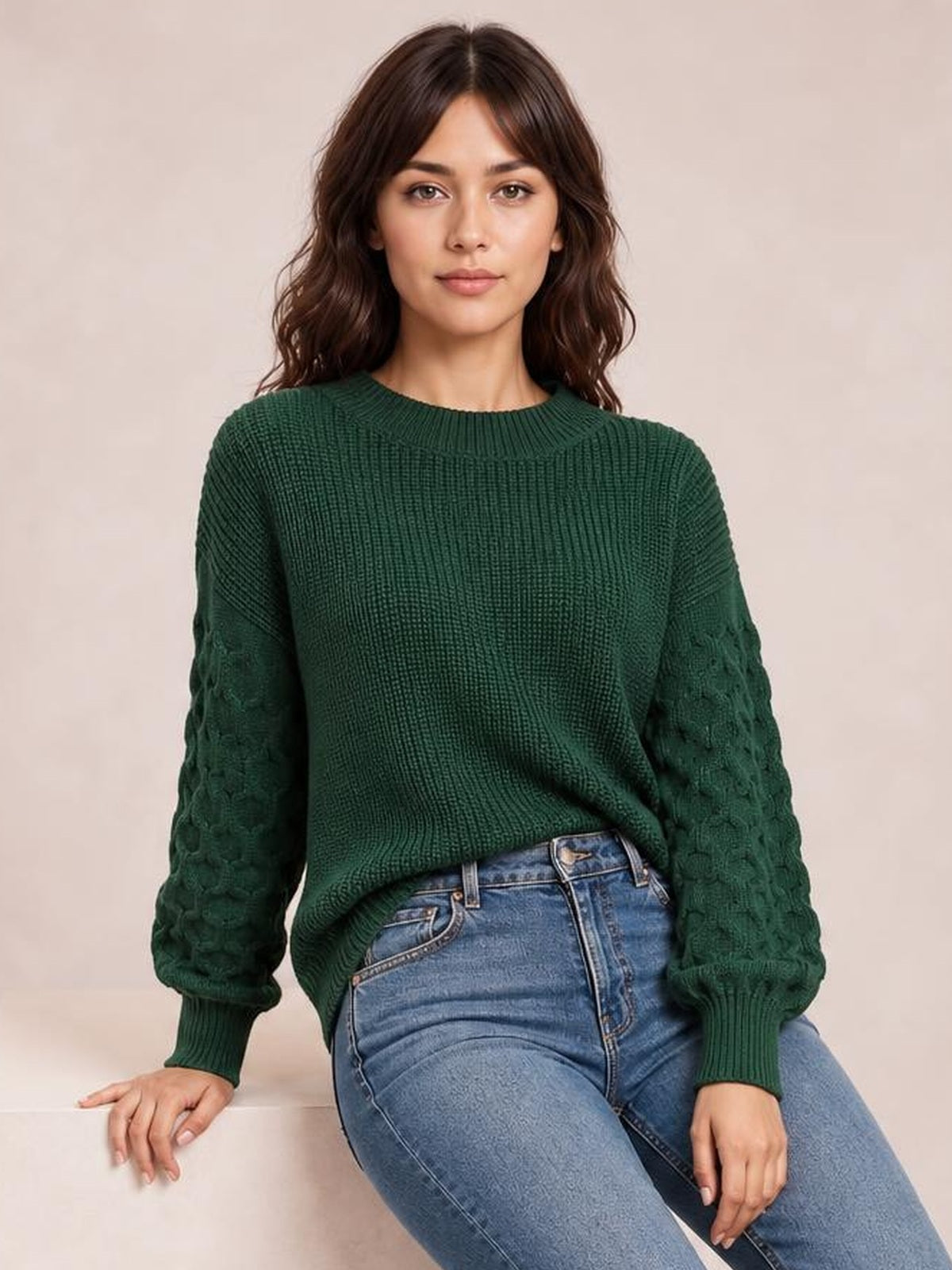 Model seated in a dark green textured sleeve round neck sweater with a ribbed neckline, cuffs, hem, relaxed silhouette, and patterned knit sleeves.