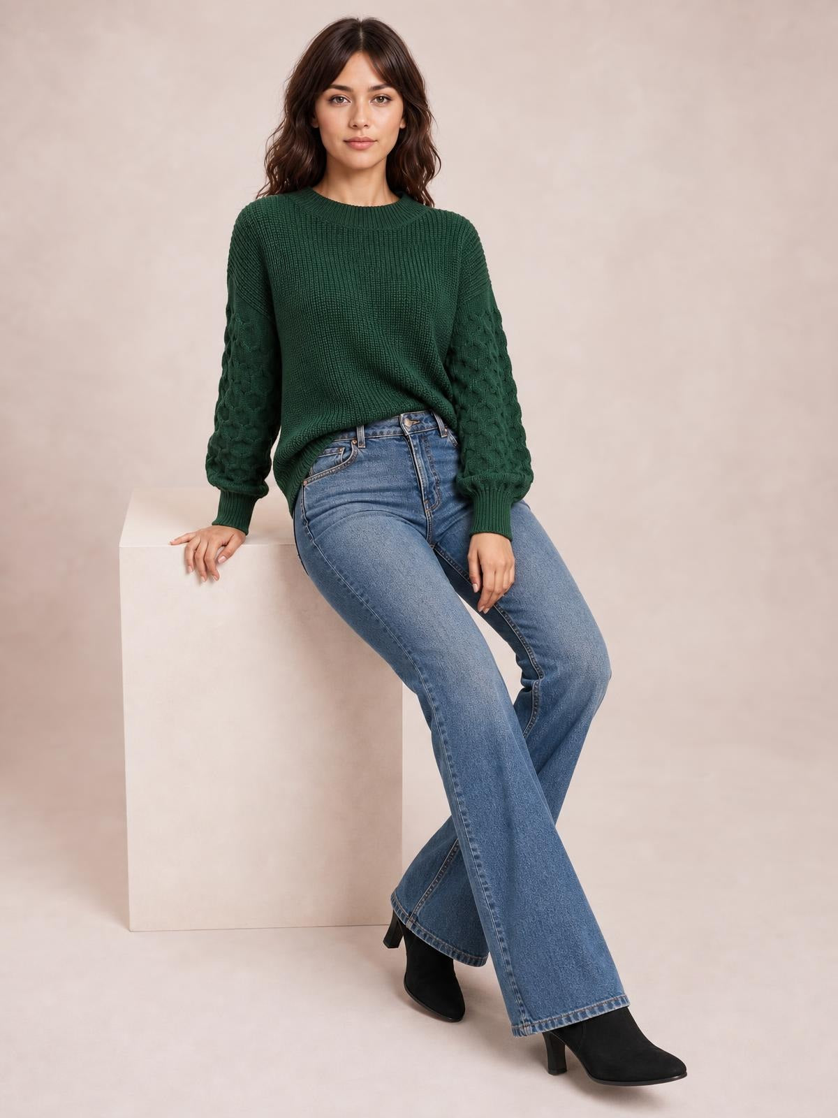 Model seated in a dark green textured sleeve round neck sweater with ribbed cuffs and neckline, relaxed silhouette, and cable-knit sleeves.