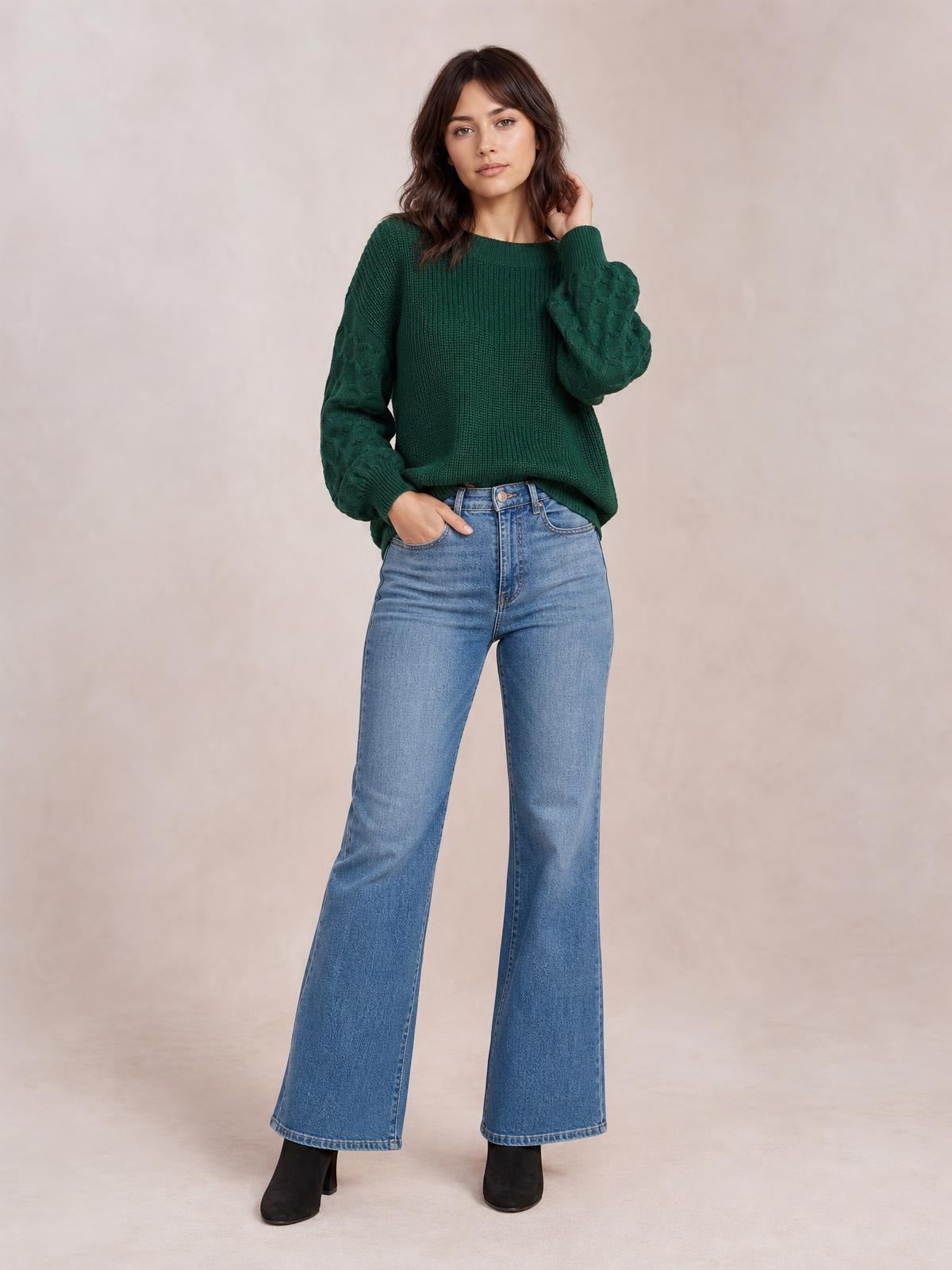 A model poses in a dark green ribbed round-neck sweater with textured cable-knit sleeves and a relaxed silhouette, one hand resting in her jeans pocket.