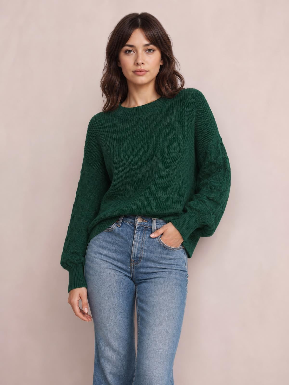 Model wears a dark green round neck sweater with textured sleeves, ribbed cuffs and hem, a relaxed silhouette, and one hand tucked into her jeans pocket.