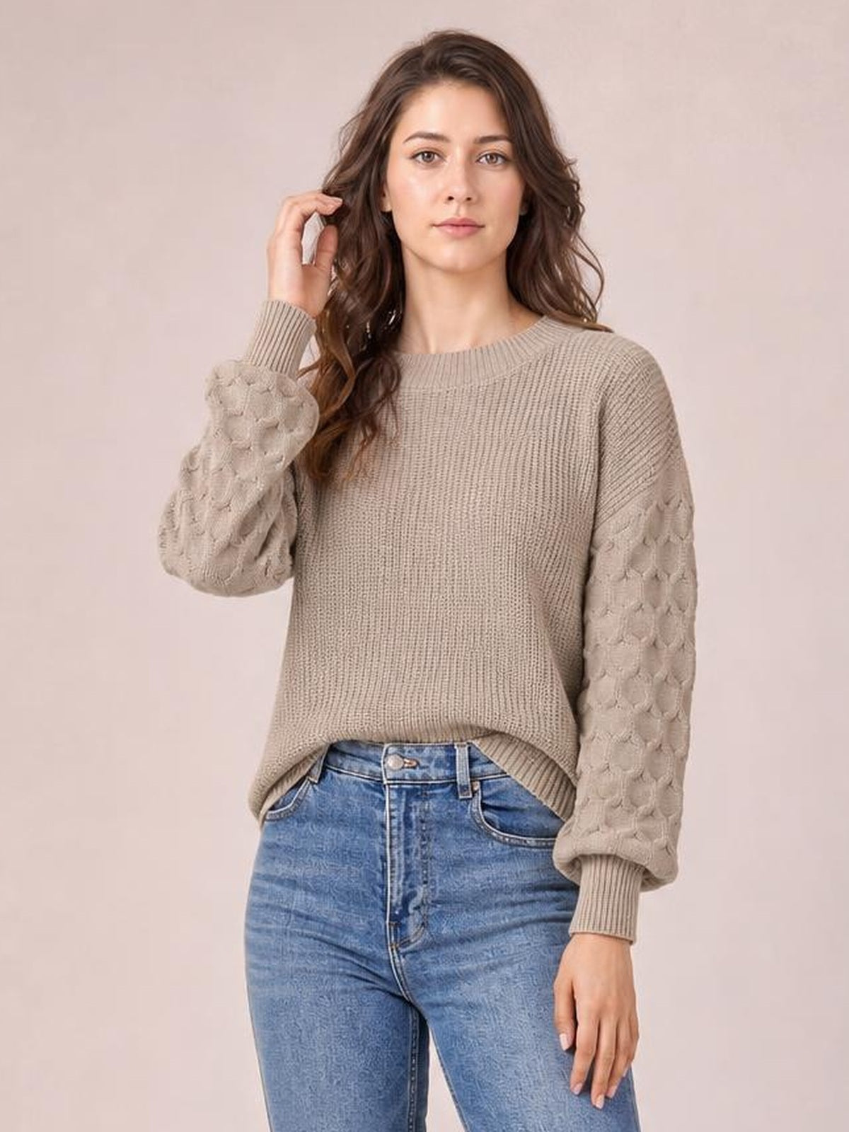 A model poses in a taupe textured sleeve round neck sweater with a ribbed body, raised patterned sleeves, relaxed silhouette, and ribbed cuffs and hem.