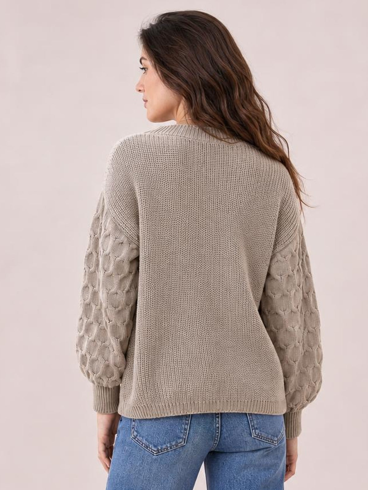 A model faces away wearing a taupe round neck sweater with textured patterned sleeves, ribbed cuffs and hem, and a relaxed, slightly oversized silhouette.