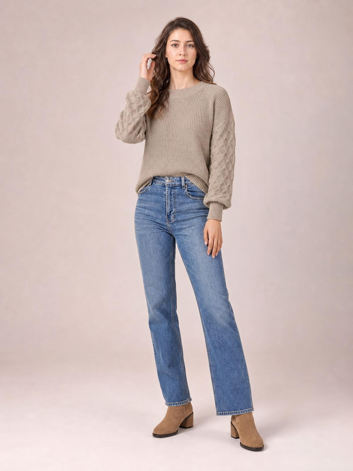 A model poses with one hand in her hair wearing a taupe round-neck sweater with a ribbed body, textured sleeves, and a relaxed silhouette.