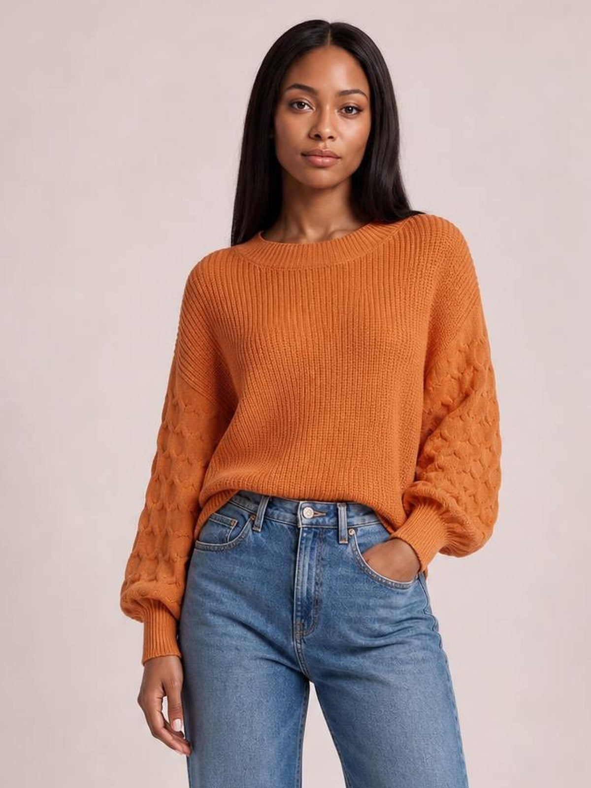 Model wearing an orange textured sleeve round neck sweater with a ribbed relaxed silhouette, patterned sleeves, and ribbed cuffs, paired with jeans.