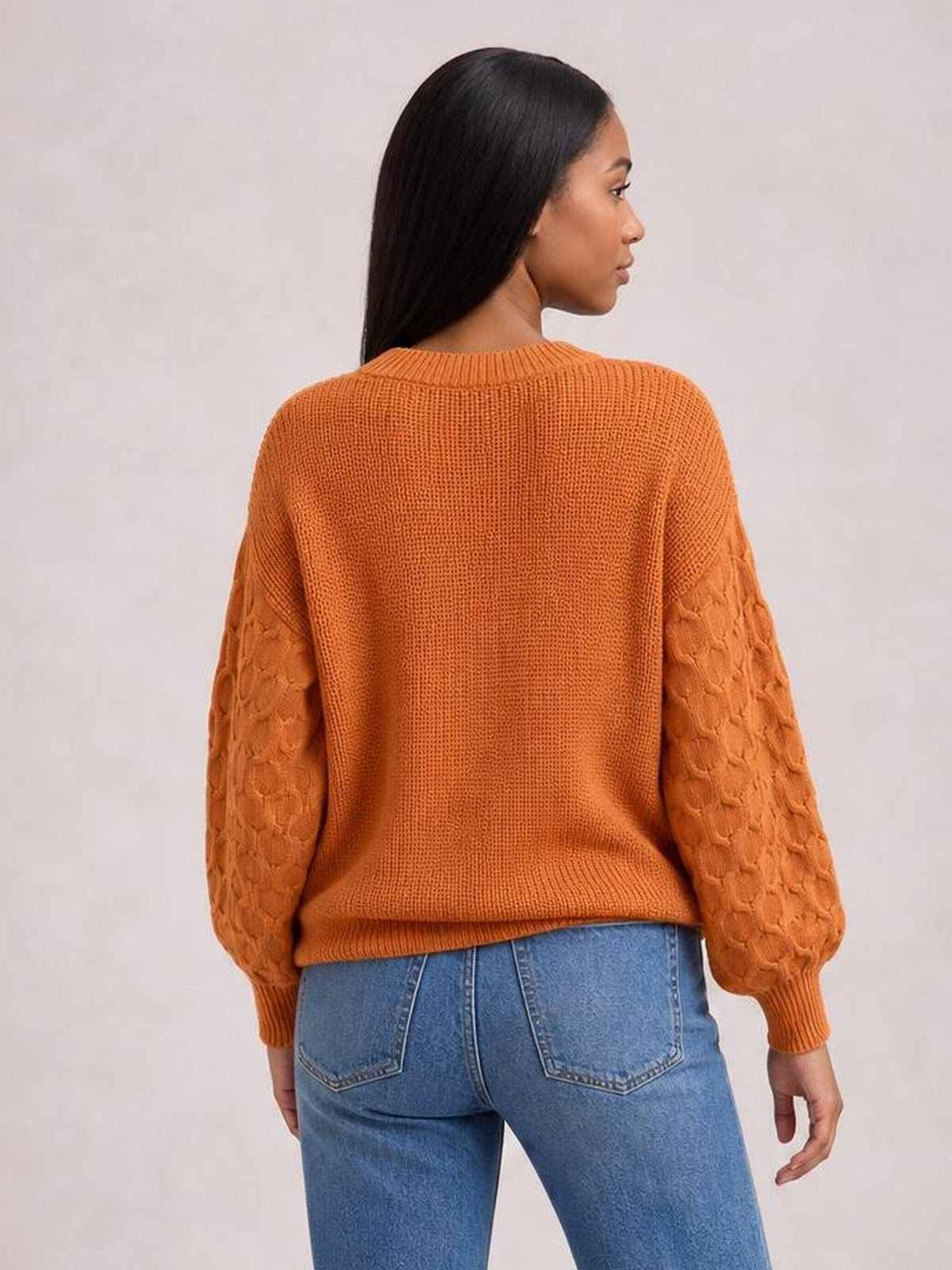 A model faces away wearing an orange textured sleeve round neck sweater with a relaxed silhouette, ribbed neckline, cuffs, and hem.