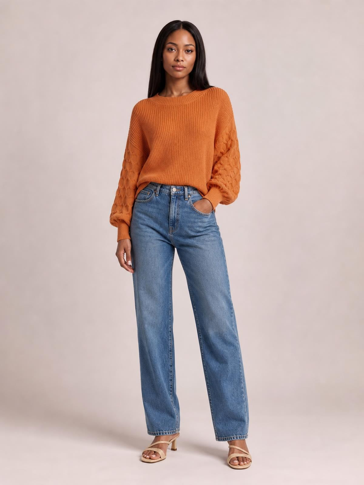 A model wears an orange textured sleeve round neck sweater with ribbed trim and a relaxed silhouette, standing with one hand in her jeans pocket.