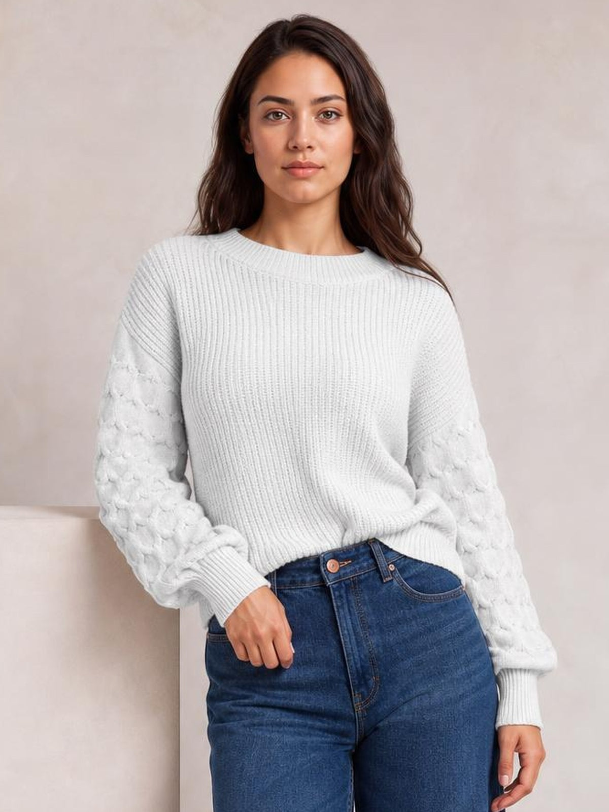 A woman wears a white round-neck sweater with a ribbed relaxed silhouette, textured sleeves, and ribbed cuffs, posing with one hand in her jeans pocket.