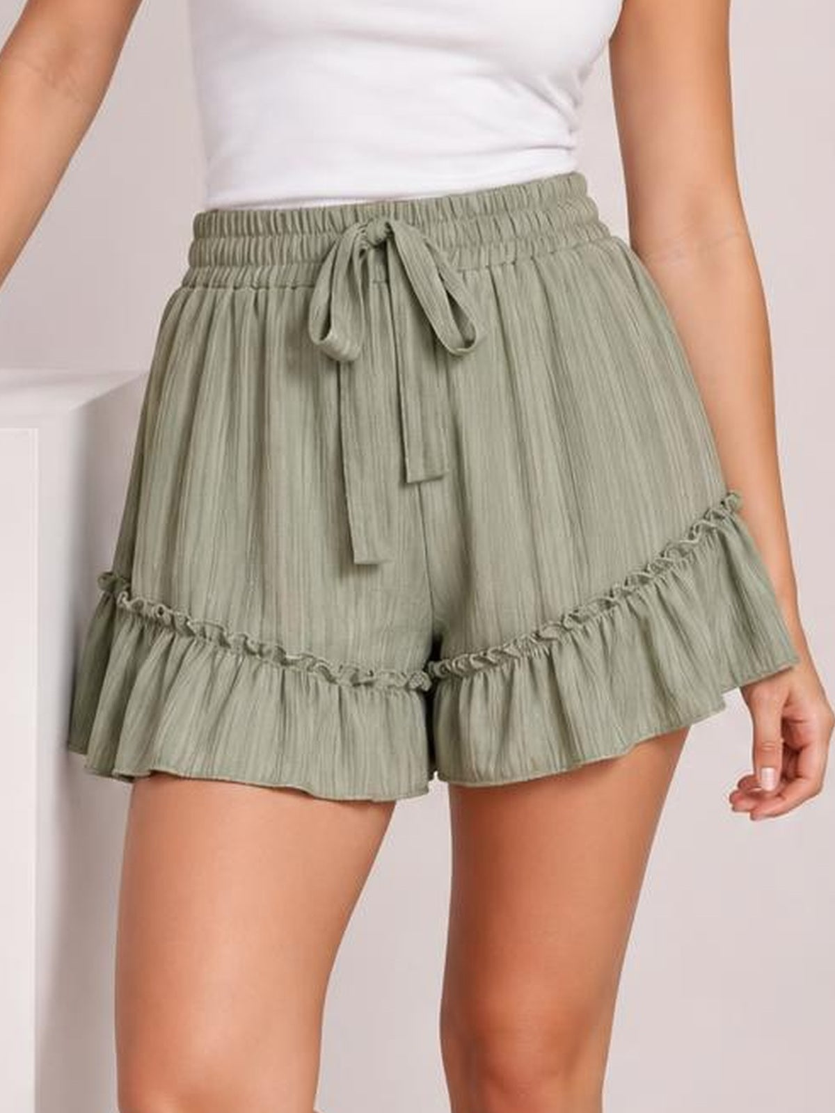 Model wearing sage green textured shorts with a smocked tie waist, relaxed silhouette, and tiered ruffle hems, shown standing with one hand at her side.